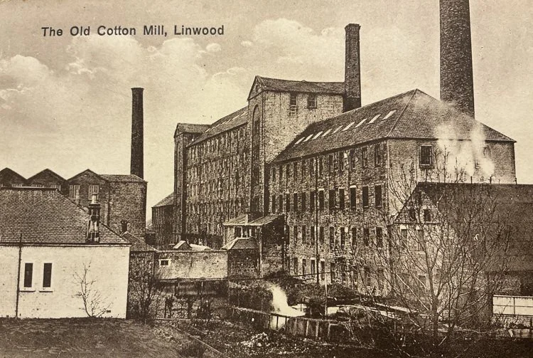A vintage black-and-white photo of the old cotton mill in Linwood, showing large brick buildings with tall chimneys, smoke rising from the factory, and a small waterway in the foreground.