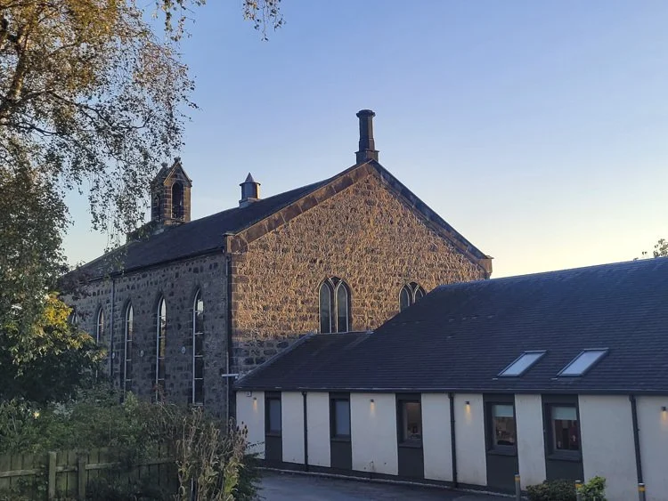 A stone church with arched windows and a small bell tower, adjacent to a modern white church hall with multiple skylights, under a clear blue sky.