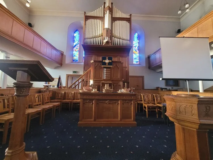 Interior of a church sanctuary with wooden pews, a central altar, a large pipe organ above the altar, stained glass windows, and a projection screen on the right.