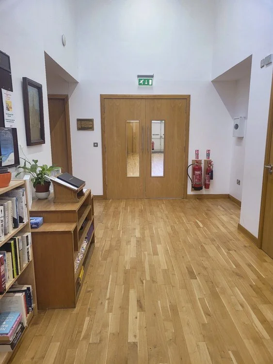 An indoor hallway leading to church hall with wooden flooring, a closed double wooden door with windows, a small bookshelf, a potted plant, fire extinguishers, and emergency exit signs.