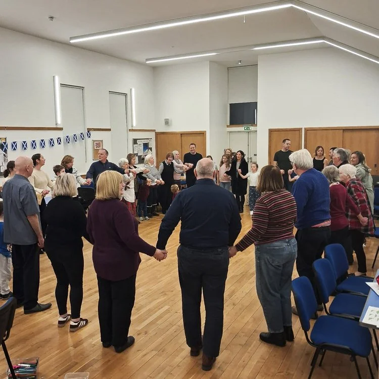 People holding hands and forming a circle in a church hall, about to sing Auld Lang Syne.