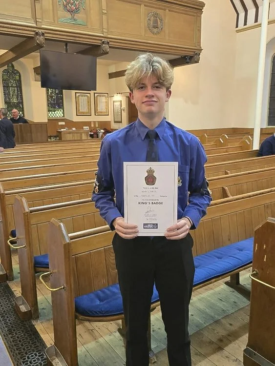 A young man dressed in a blue Boys' Brigade uniform holding a certificate that reads 'King's Badge' inside a church with wooden pews and stained glass windows.