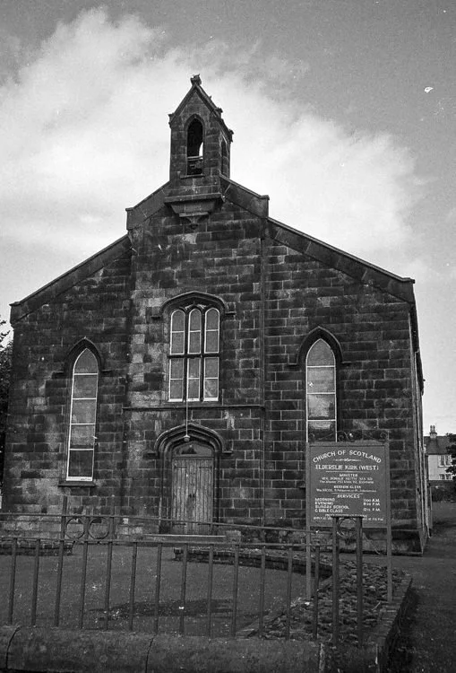 A black-and-white photo of an old stone church with Gothic-style arched windows and a wooden door, surrounded by a metal fence. There is a sign outside indicating it is the Church of Scotland in Elder Street.