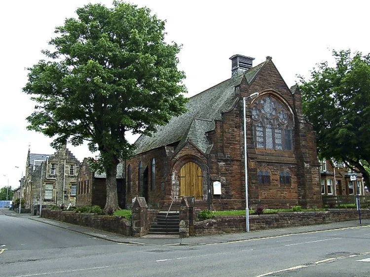 A historic red sandstone church with stained glass windows, a wooden door, and stone steps, surrounded by trees and located at a street corner.
