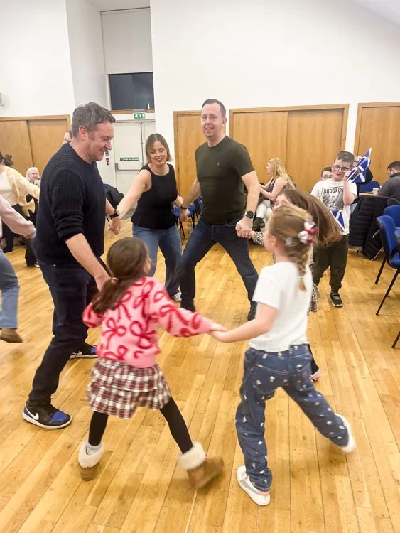 People of different ages dancing and holding hands in a circle in a church hall with wooden floors and chairs.