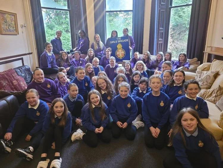 Group of young girls in purple and blue uniforms sitting and standing inside a room with large windows, holding a sign that reads 'The Girls Brigade'.