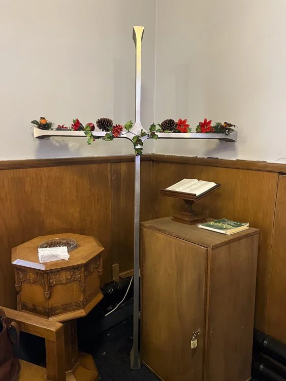 A large metal cross from Linwood decorated with poinsettia flowers, pinecones, and greenery, standing in a corner of a church with wooden paneling, near a small wooden cabinet with an open Bible and a smaller closed book on top.