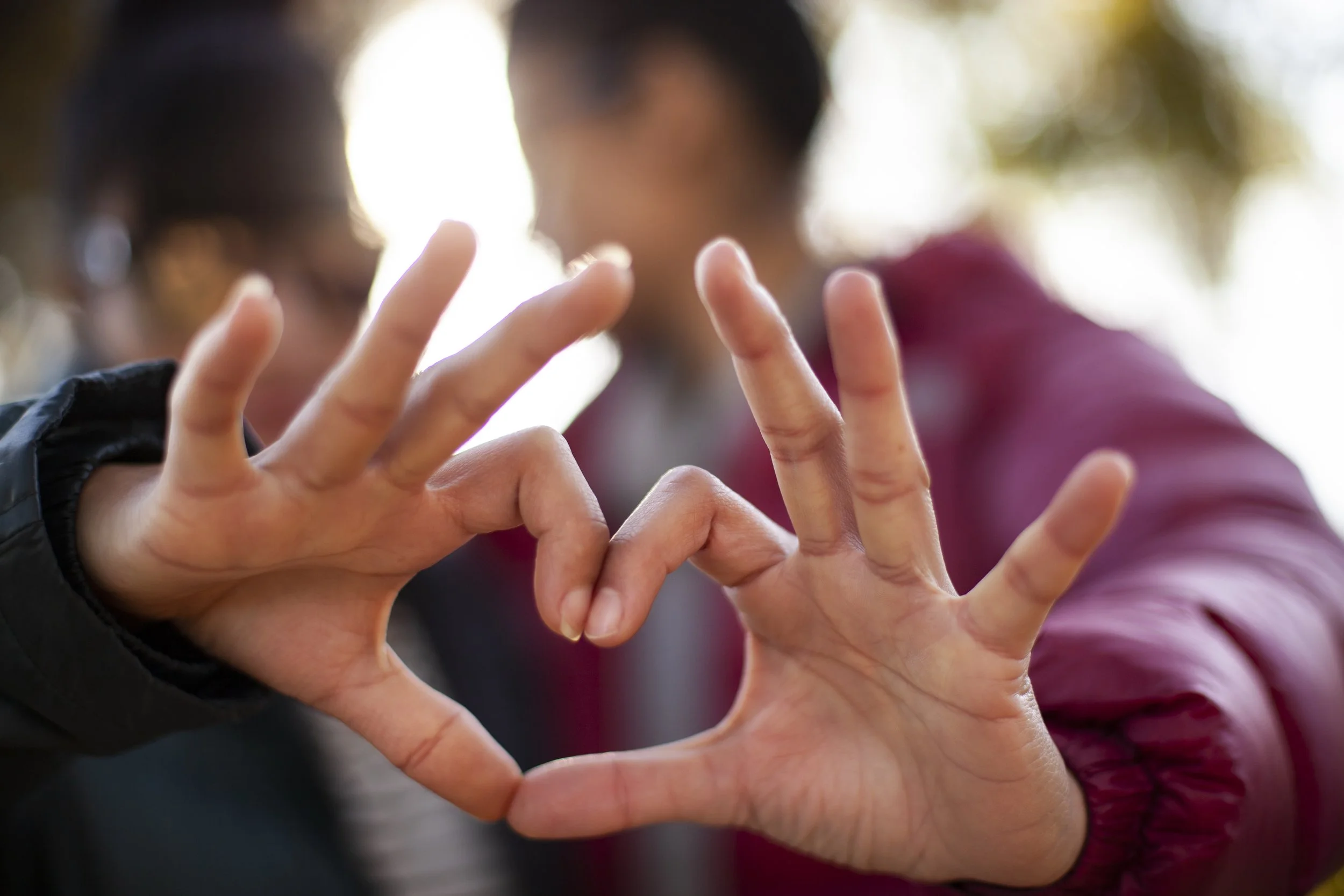 Close up of 2 hands from 2 people held together to make the shape of a heart. The people are blurred in the background and appear to be looking at each other.