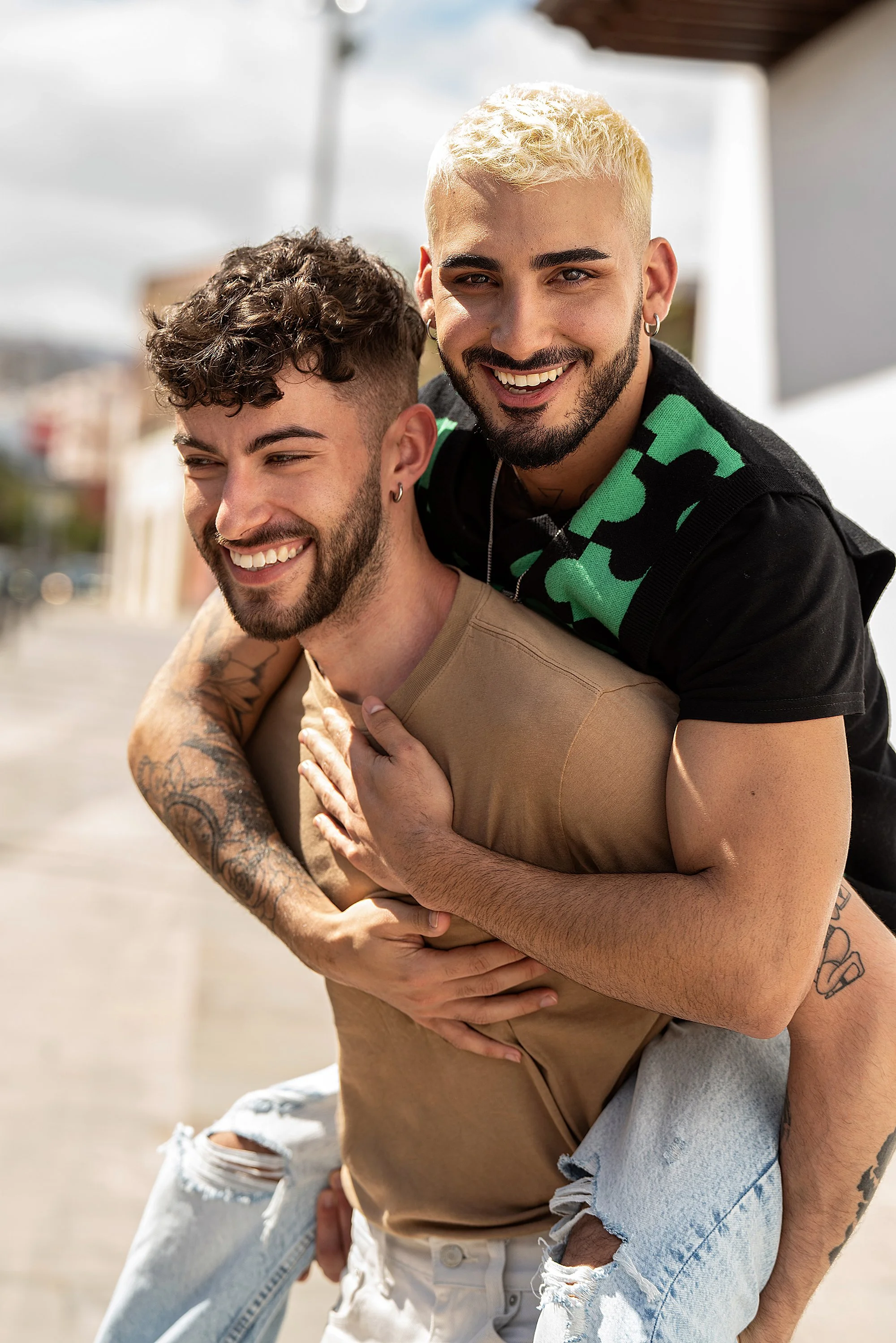 A younger man with dark hair gives a piggy back ride to another younger man with bleach blonde hair. They are smiling and laughing.