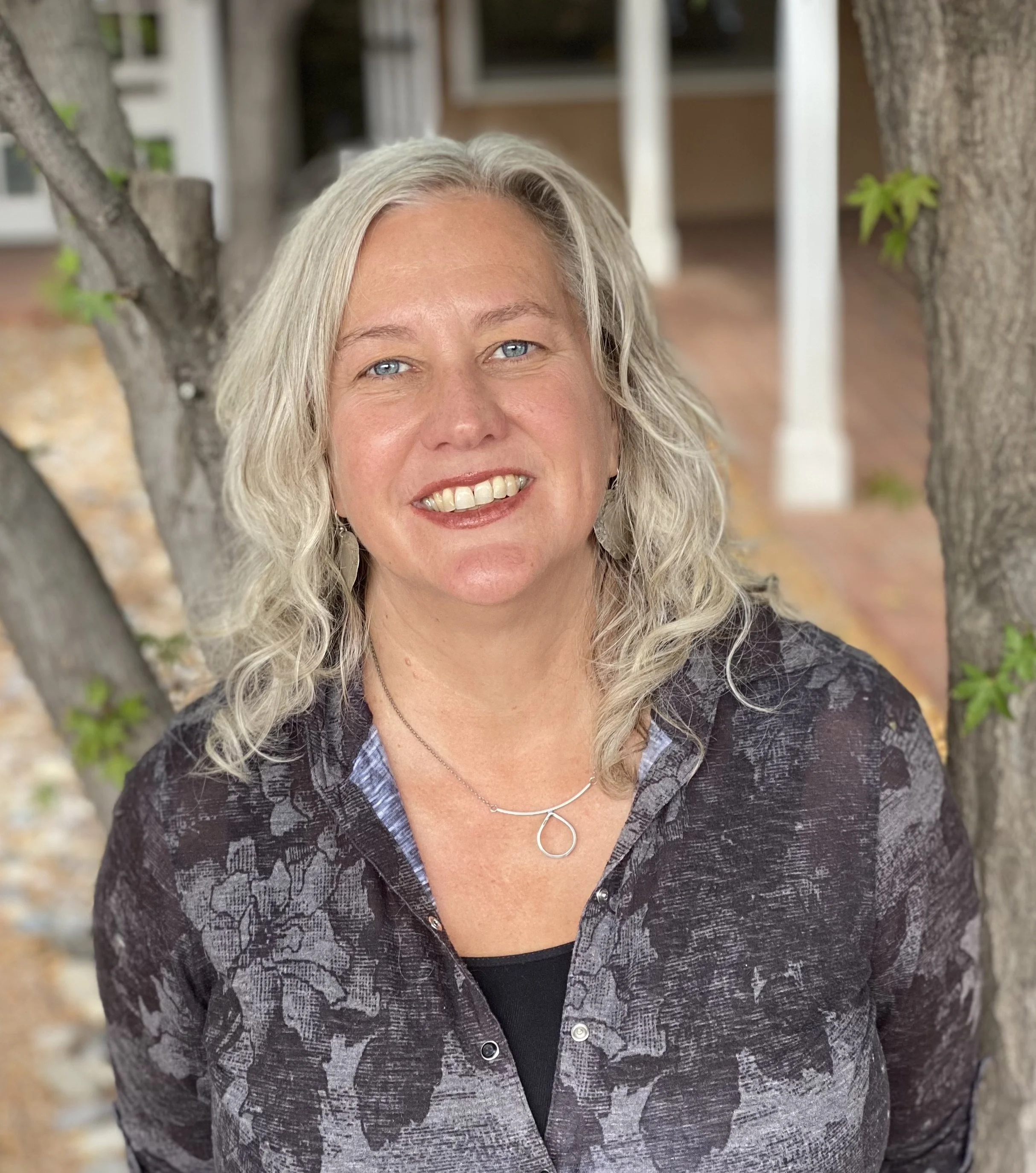 Wendy Johnson, a woman with shoulder length gray hair, stands amongst trees and smiles up at us.
