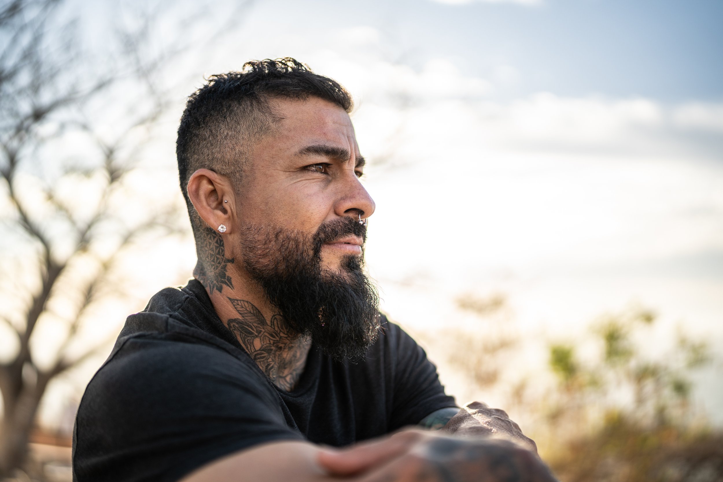 Latino man with a long beard, septum piercing, and many tattoos looks off into the distance to our right.