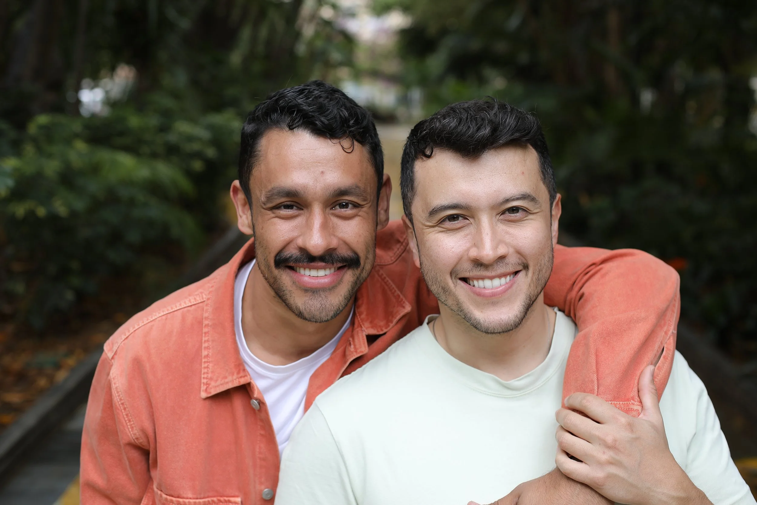 2 men with dark hair smile at us. The one on the left has his arm over the other's shoulder, while the second man holds his wrist gently.