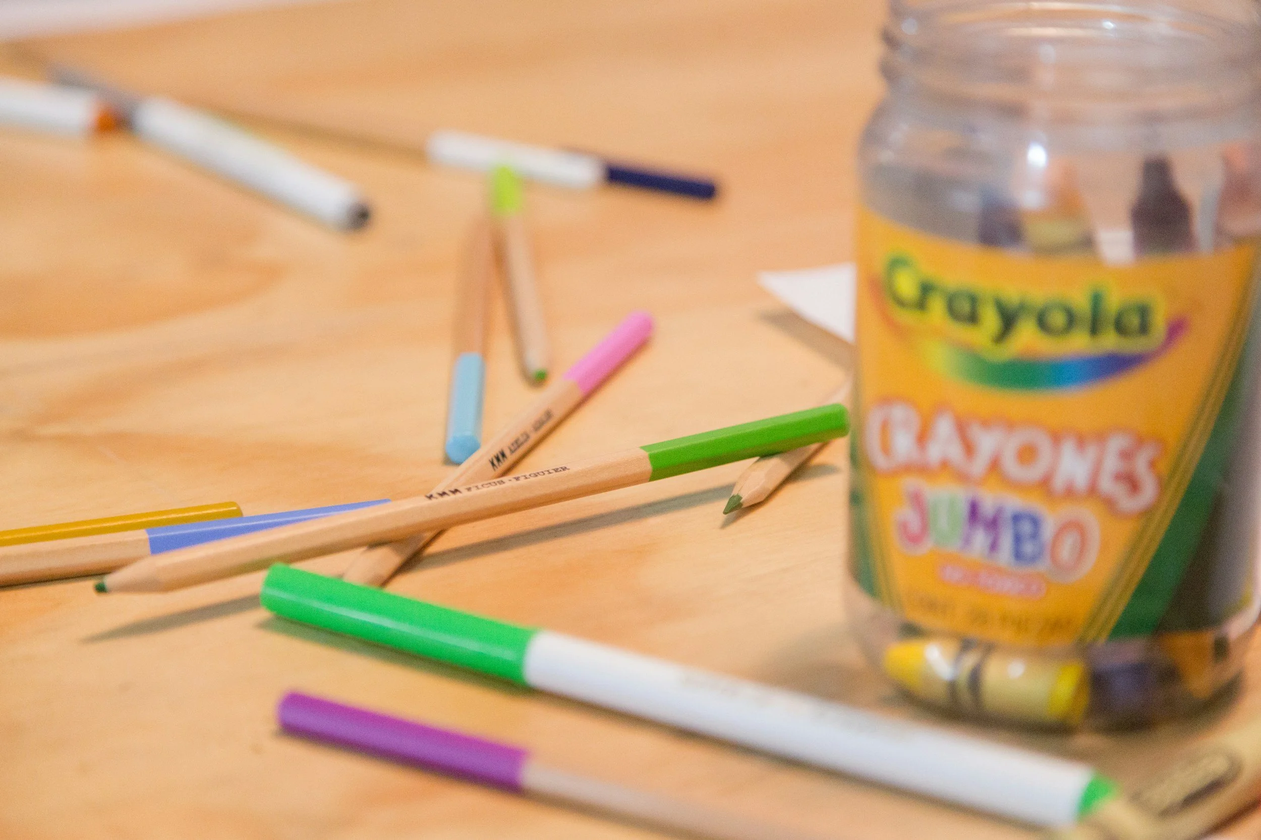 Colorful markers and crayons scattered on a wooden table with an open container of Crayola crayons in the background.