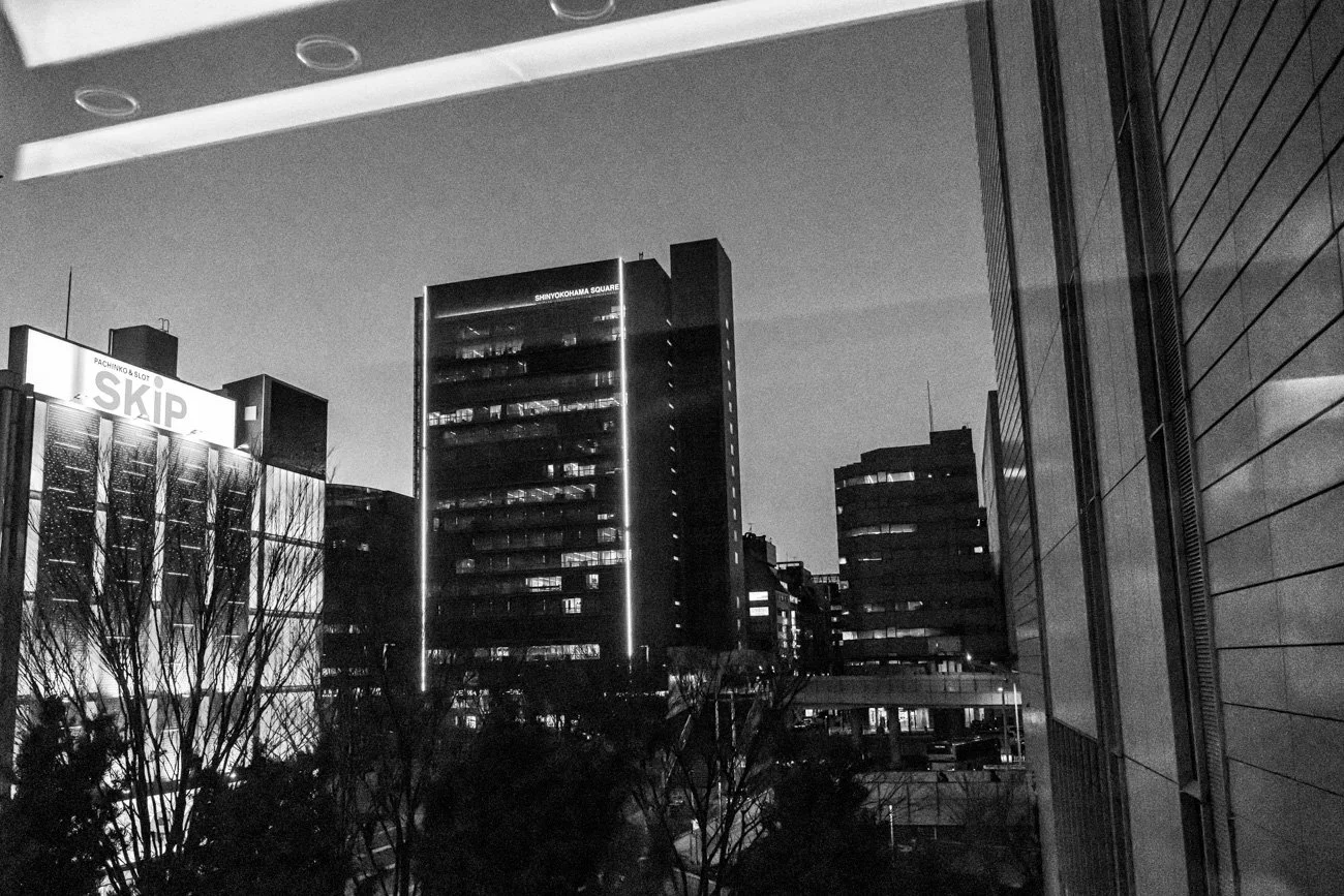 Black and white photo of city buildings at night, with signs for 'SKIP' and 'SHIN-YOKOHAMA SQUARE' visible, and trees in the foreground.
