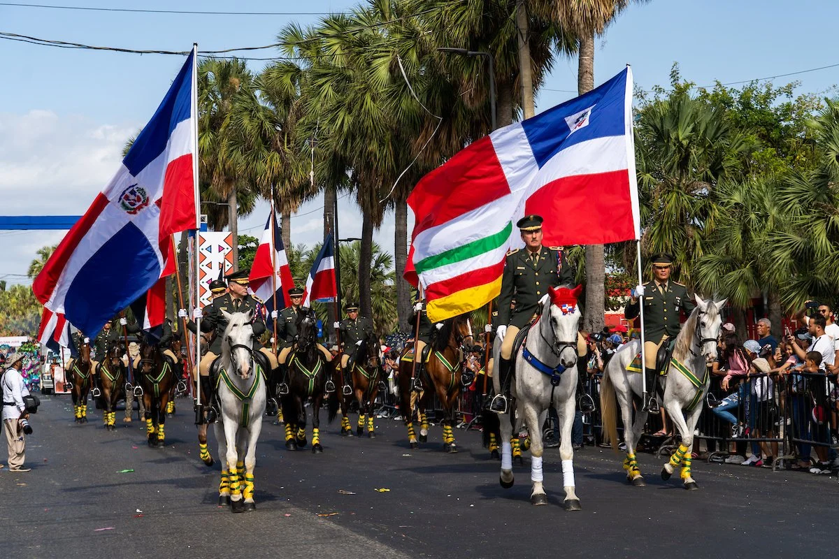 Military parade with soldiers on horseback carrying Dominican, Guatemalan, and other flags, with spectators and palm trees in the background.
