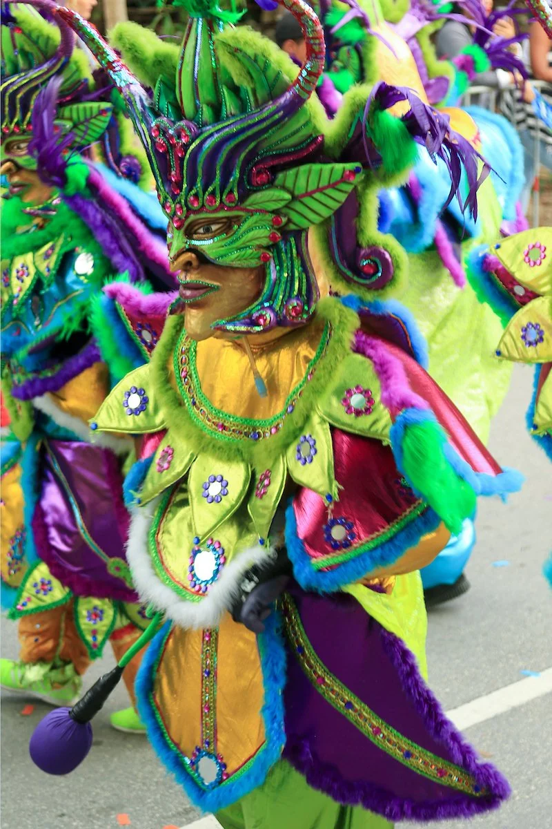 Person dressed in a colorful, elaborate costume with a large, feathered and jeweled mask, participating in a parade or carnival.