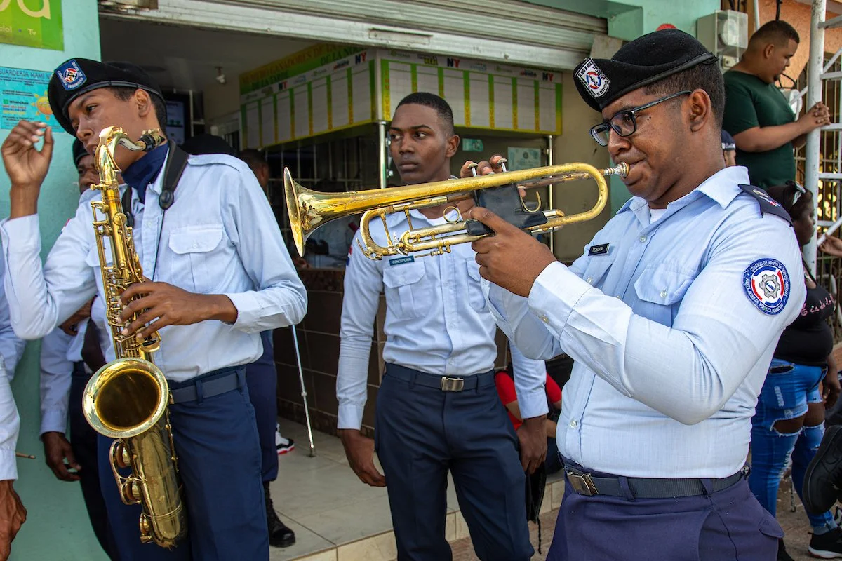 Three police officers in uniform playing musical instruments, with a woman and a young girl in the background. The officer on the left plays a saxophone, and the officer on the right plays a trumpet.