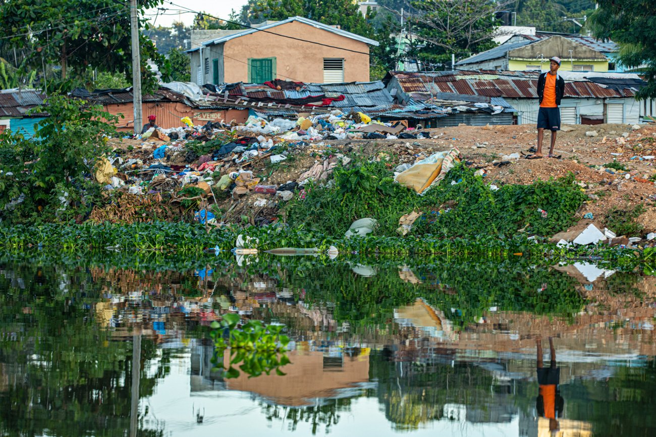 A young man stands on a dirt hill next to a pile of trash and debris near a body of water, with makeshift houses in the background.