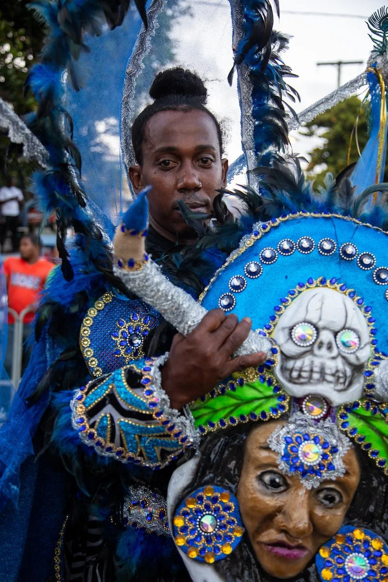 A person dressed in a colorful, elaborate costume with blue and black feathers, rhinestones, and a skull decoration, holding a decorated stick at a parade or festival.