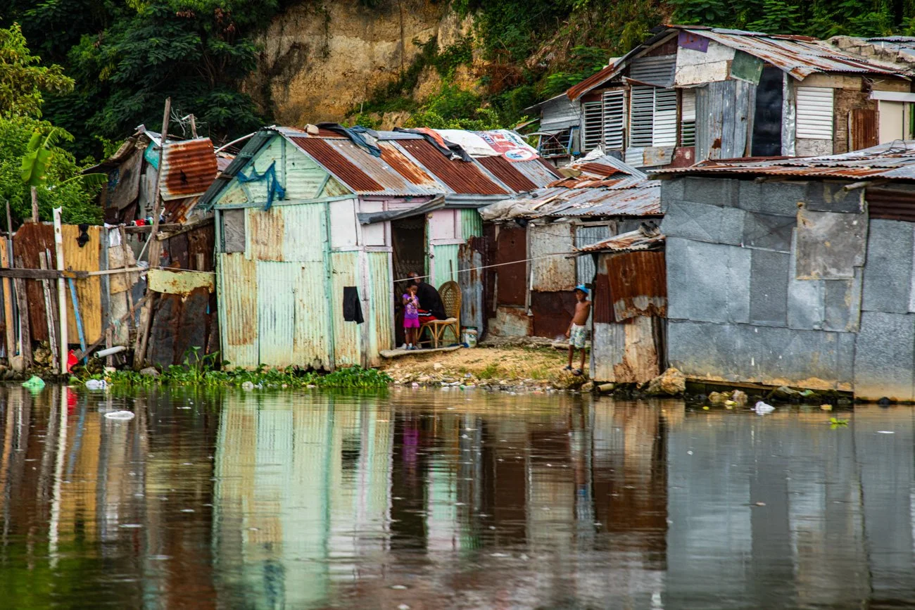 Shanty houses made of rusted metal sheets along a riverbank with two children and an adult outside the homes, surrounded by lush greenery.