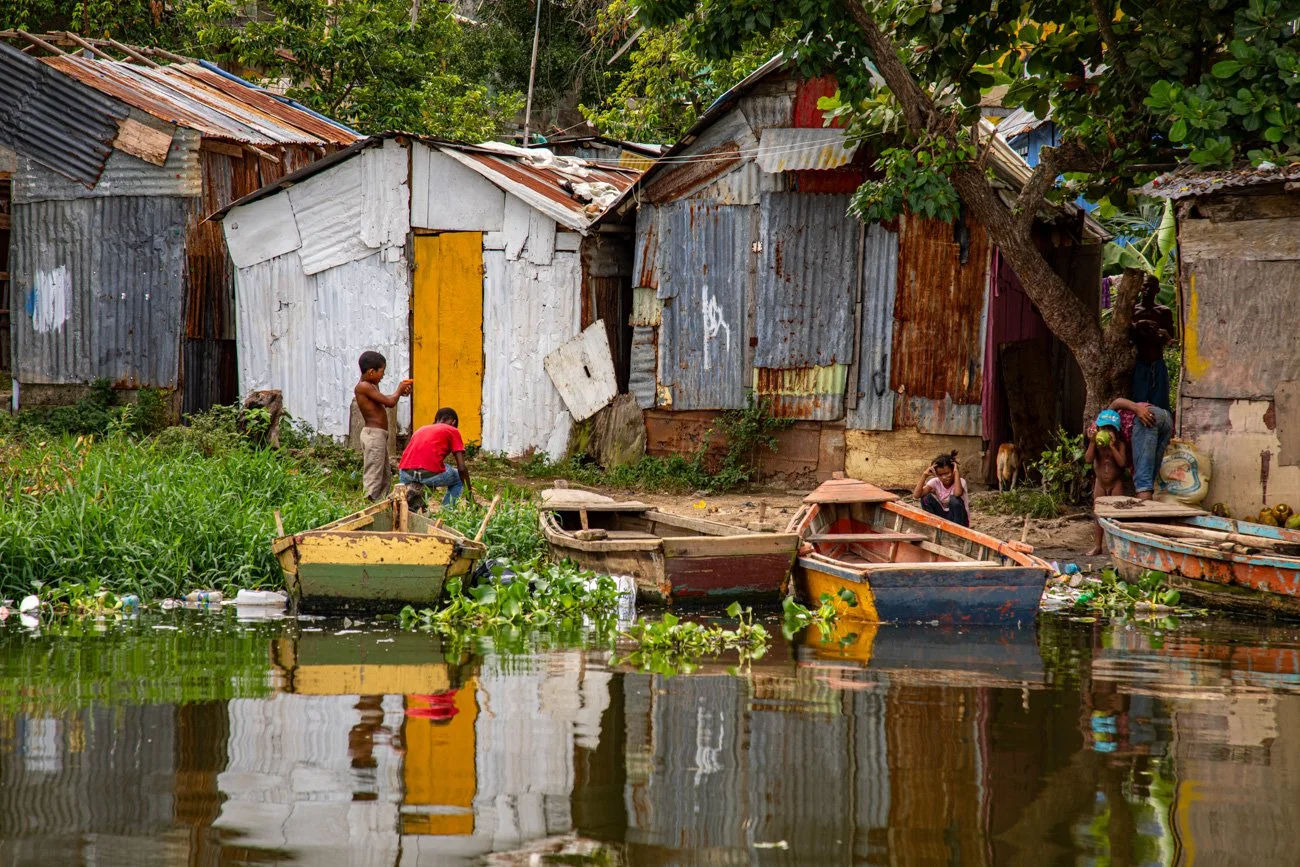 Children and adults near a river with rowboats, in front of worn, makeshift houses made of corrugated metal and wood, surrounded by trees and greenery.
