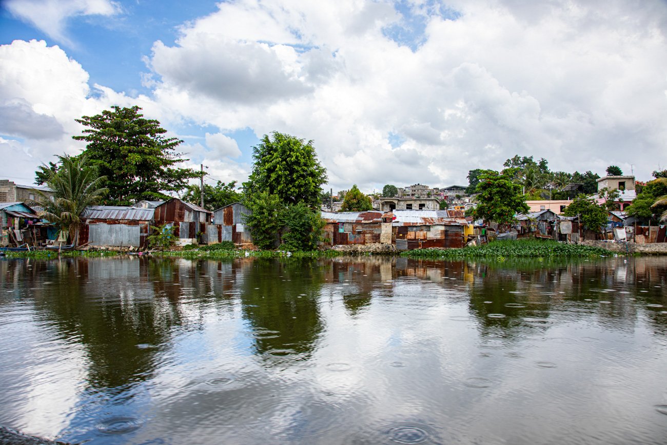 A riverside scene with shabby, rusted metal dwellings and lush green trees under a cloudy sky, reflected in the water.