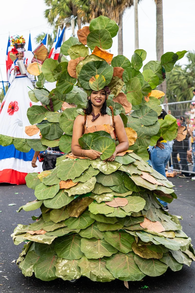 A woman dressed in a costume made of large green and brown leaves, standing in a parade with a crowd and flags in the background.