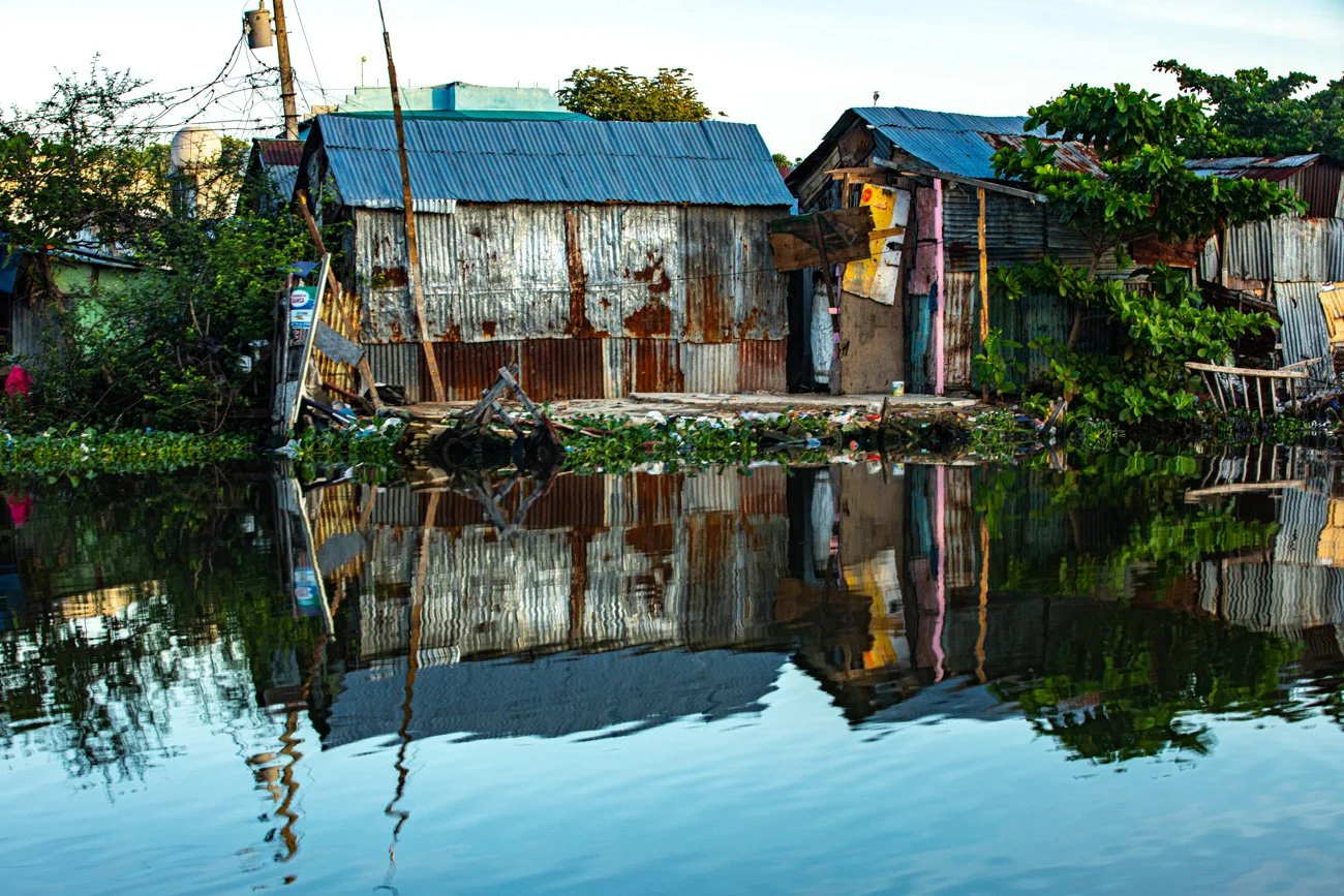 A rustic waterfront scene featuring makeshift houses with rusted metal and wooden walls, some with corrugated metal roofs, surrounded by greenery, and reflected in the calm water.