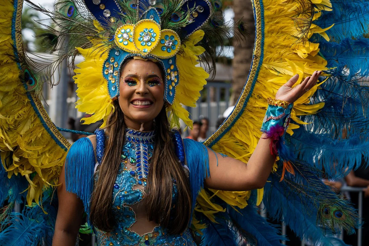 A woman in a bright yellow and blue feathered carnival costume with a headdress, smiling and waving.