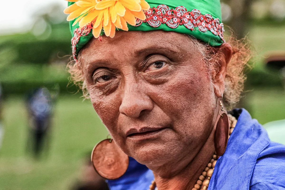 Close-up of an elderly woman with tan skin, wearing a green headscarf decorated with a yellow flower and red and white embroidery. She has large earrings, and her expression appears pensive or serious. She is outdoors with blurred greenery in the bac
