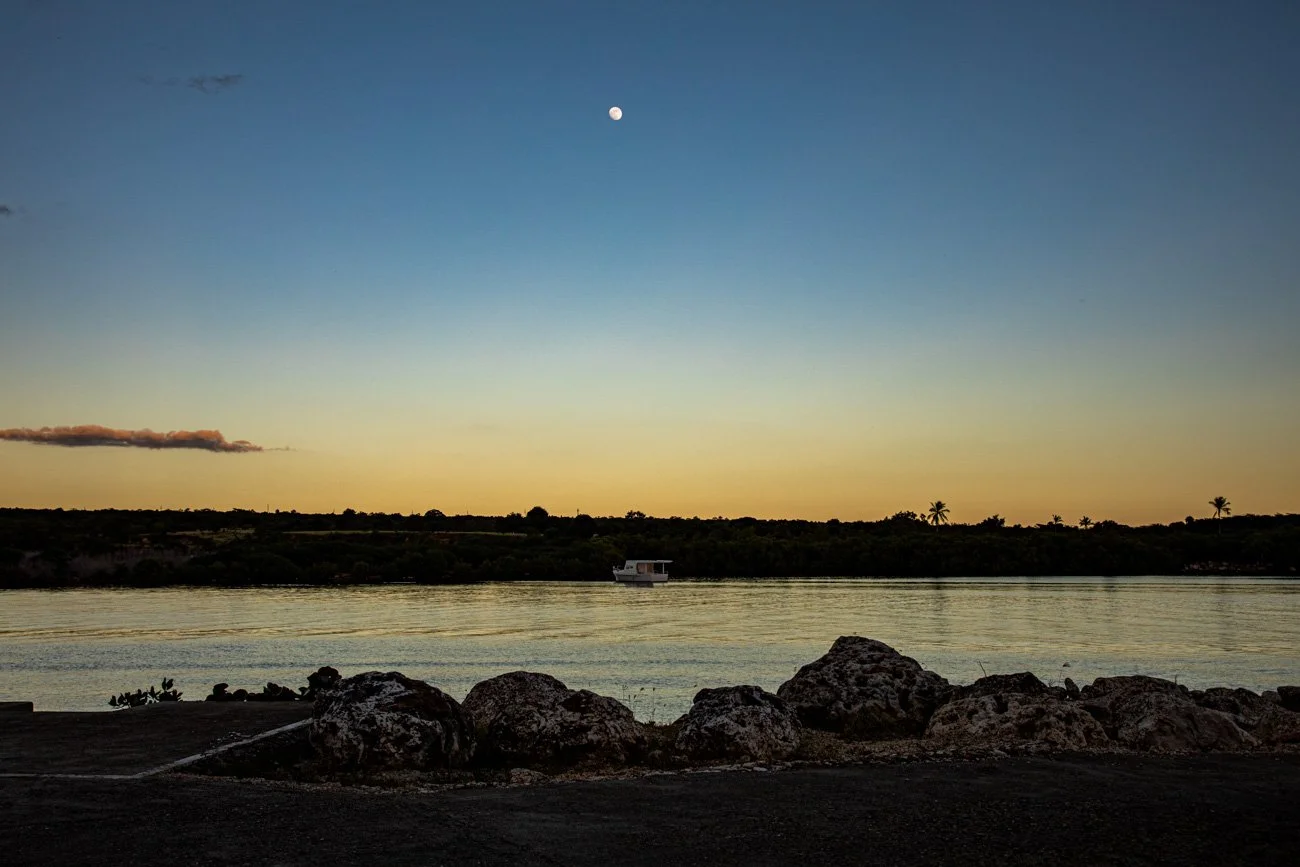 A riverside scene at sunset with a boat on the water, rocks in the foreground, and a moon in a clear sky.