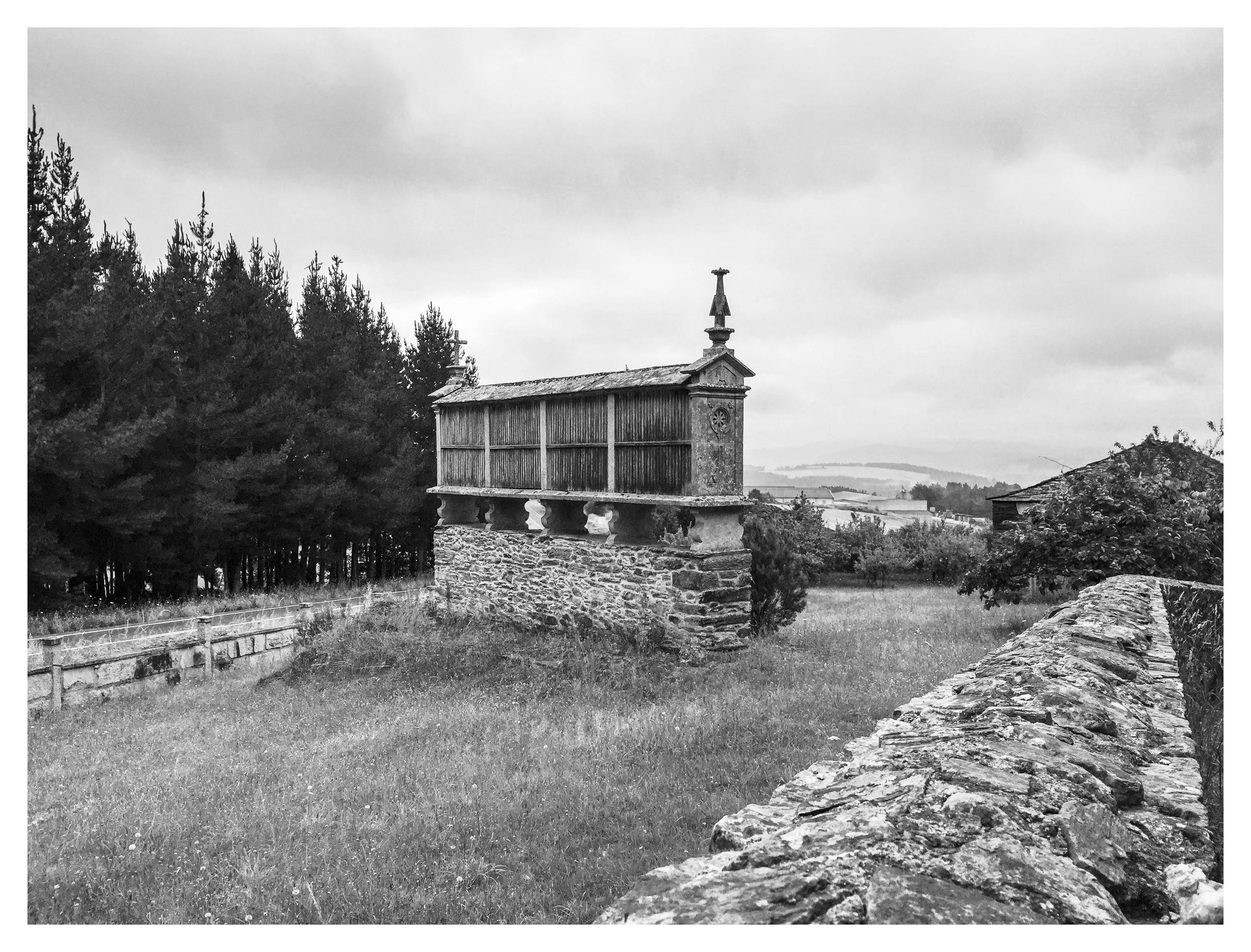 Black and white photo of a small wooden and stone chapel-like structure on a grassy field, with trees on the left and a stone wall on the right, overcast sky in the background.