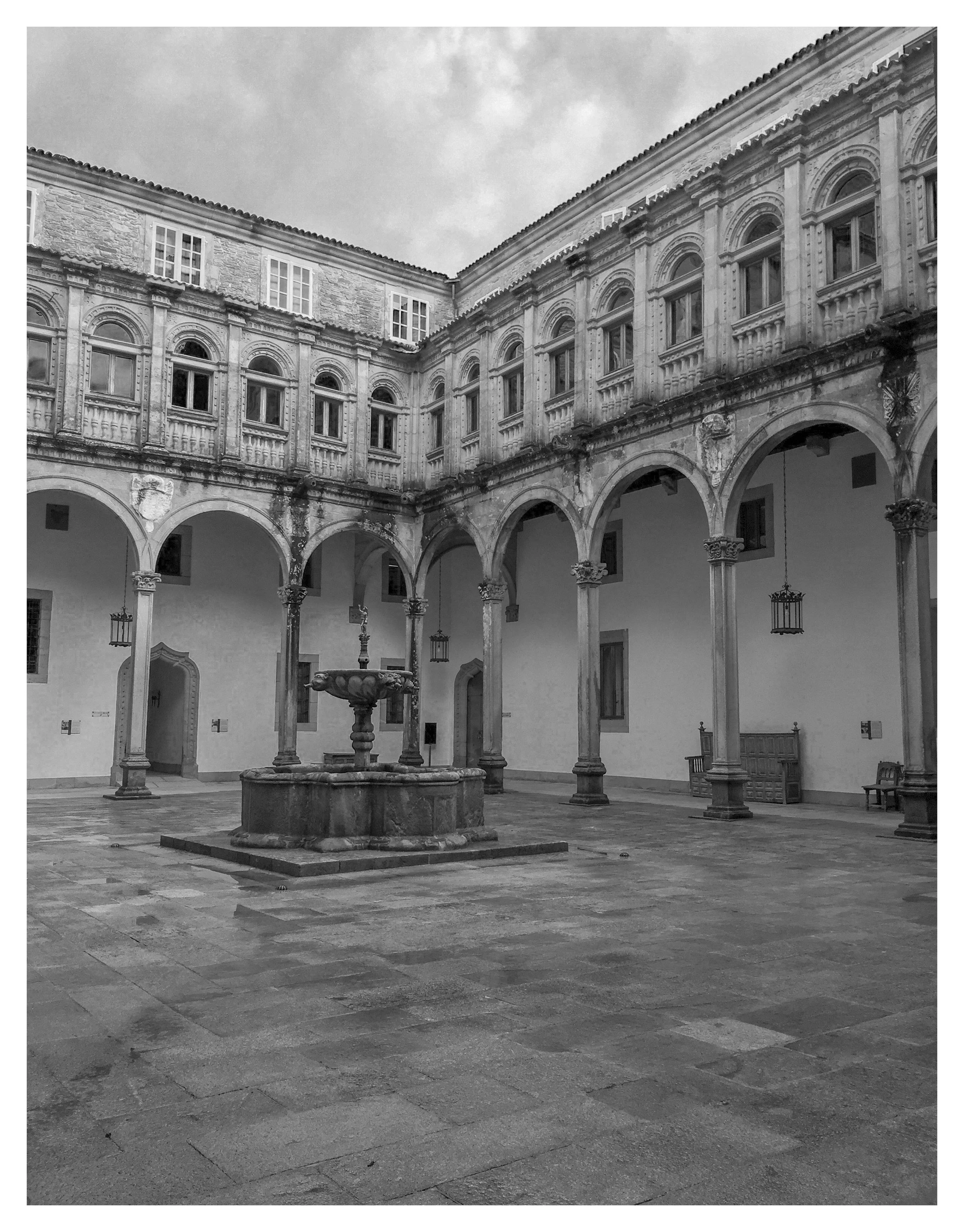 An inner courtyard of a historical building with arched columns, a central fountain, benches, and hanging lanterns, in black and white.