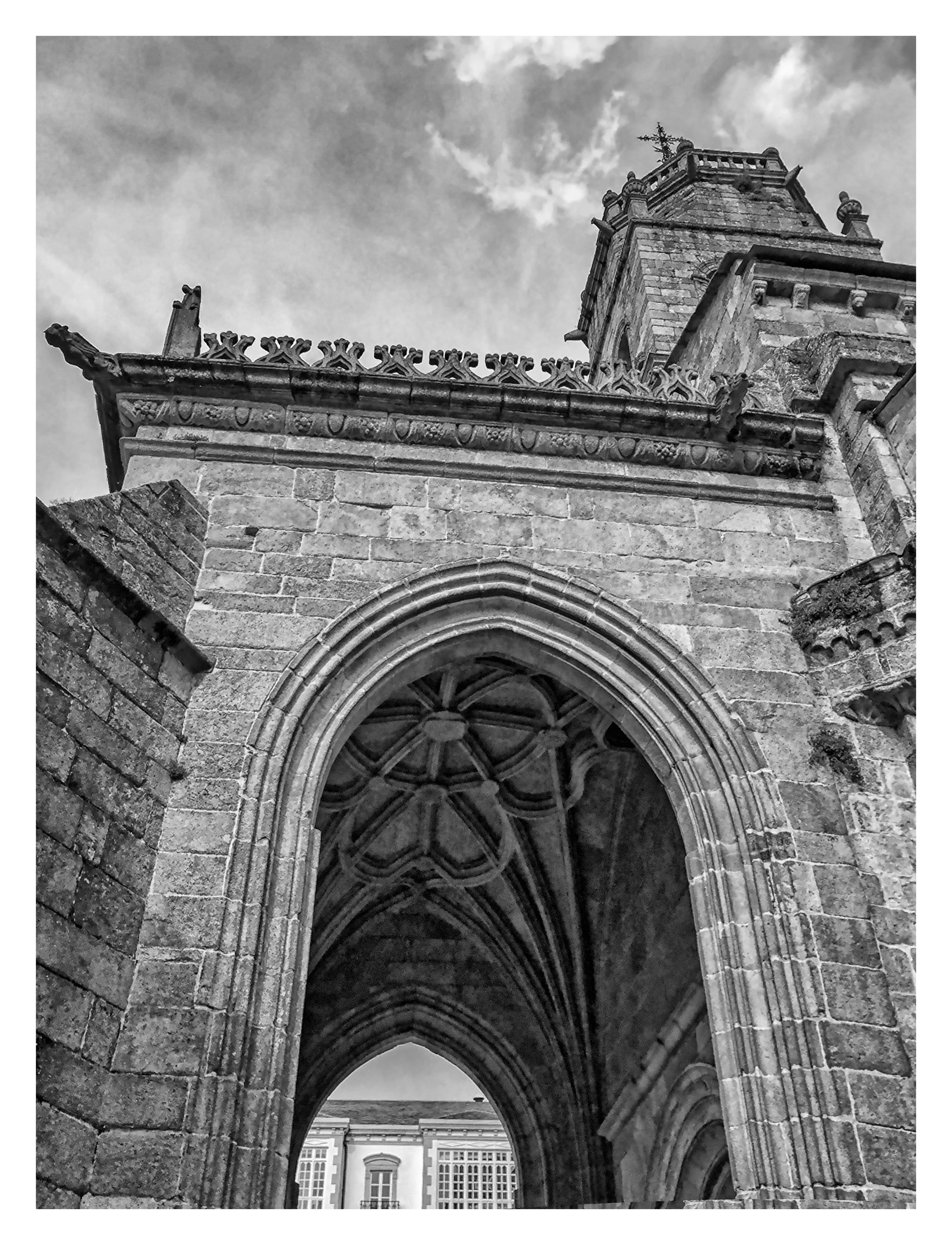 Black and white photo of a Gothic-style church archway with detailed stonework and a view of the sky and clouds above.