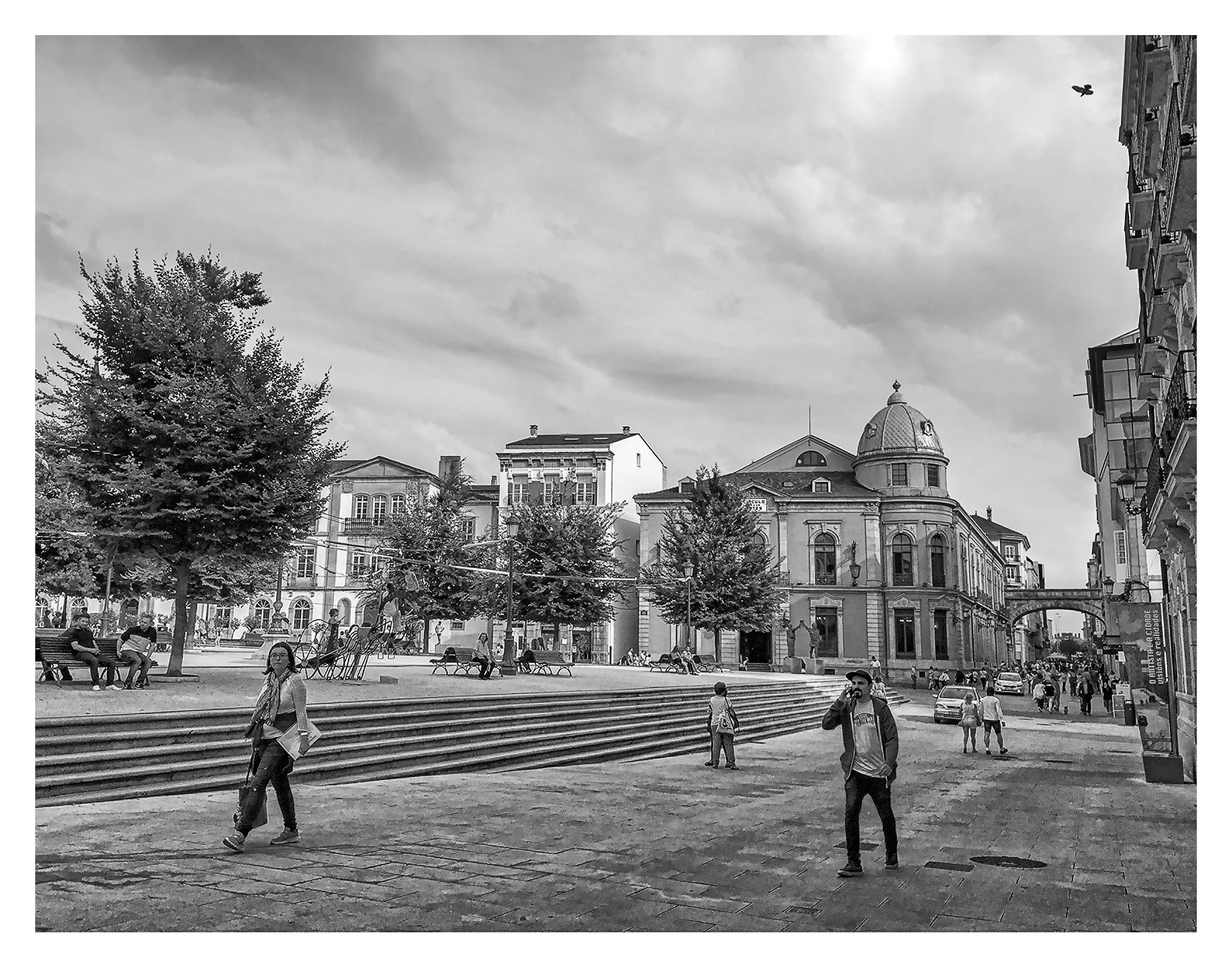 A city square with people walking, sitting on benches, and trees. There are historic buildings, including a domed structure, a bridge-like walkway, and a cloudy sky overhead.
