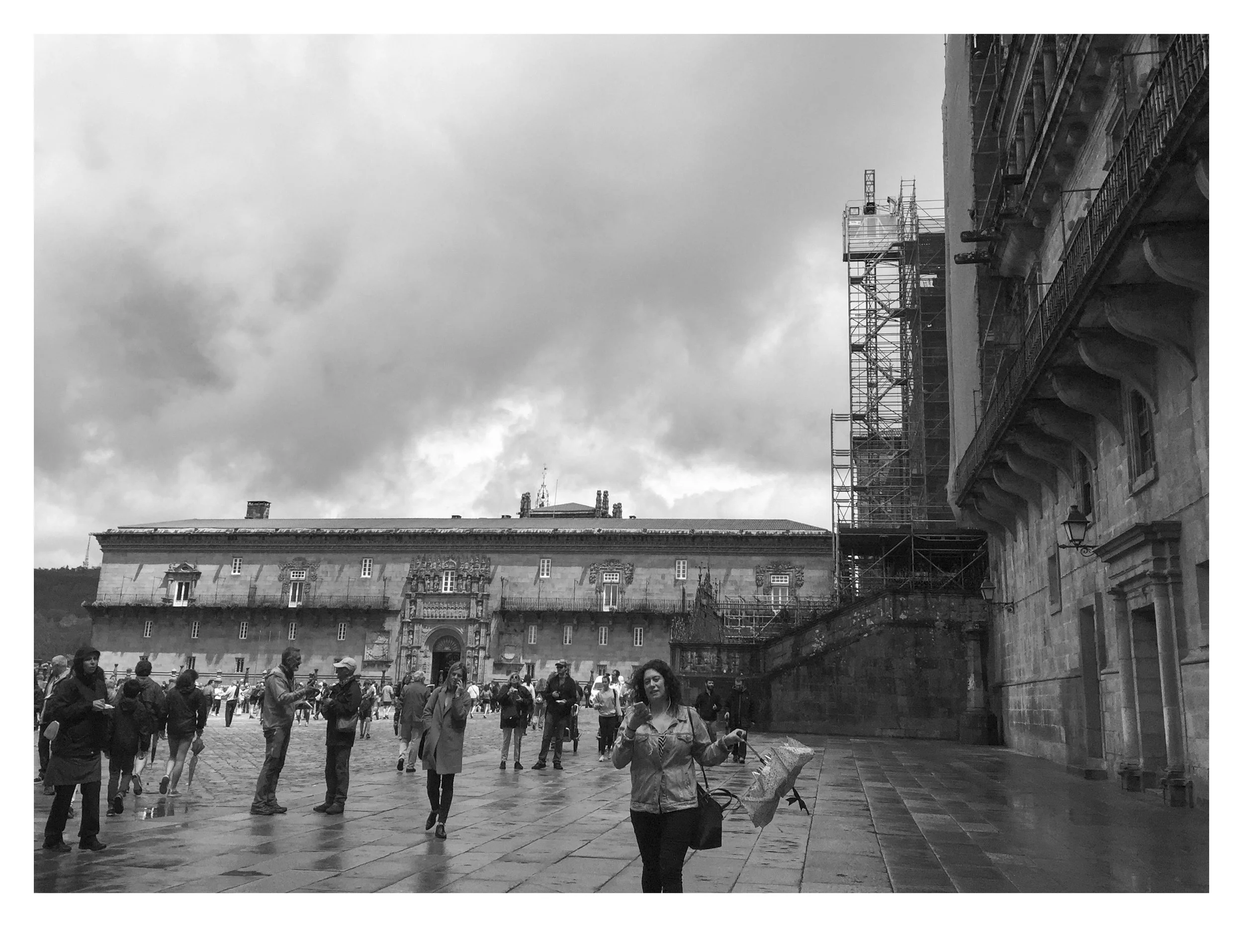 People walking and standing in a city square with historic buildings, stone paving, and scaffolding on the right side, under a cloudy sky.