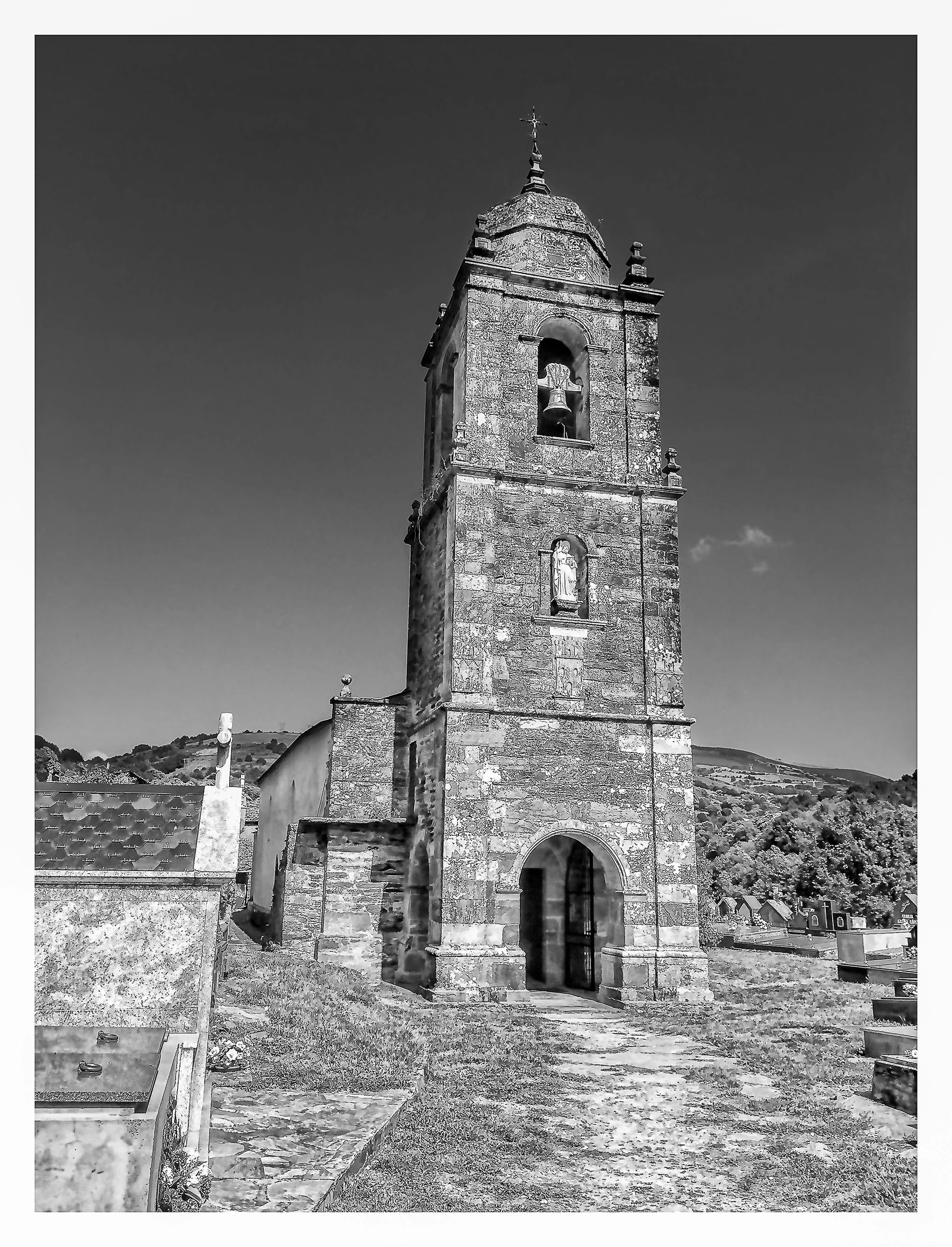 A black and white photo of a stone church bell tower with a cross on top, situated in a graveyard with headstones, against a backdrop of hills and a clear sky.