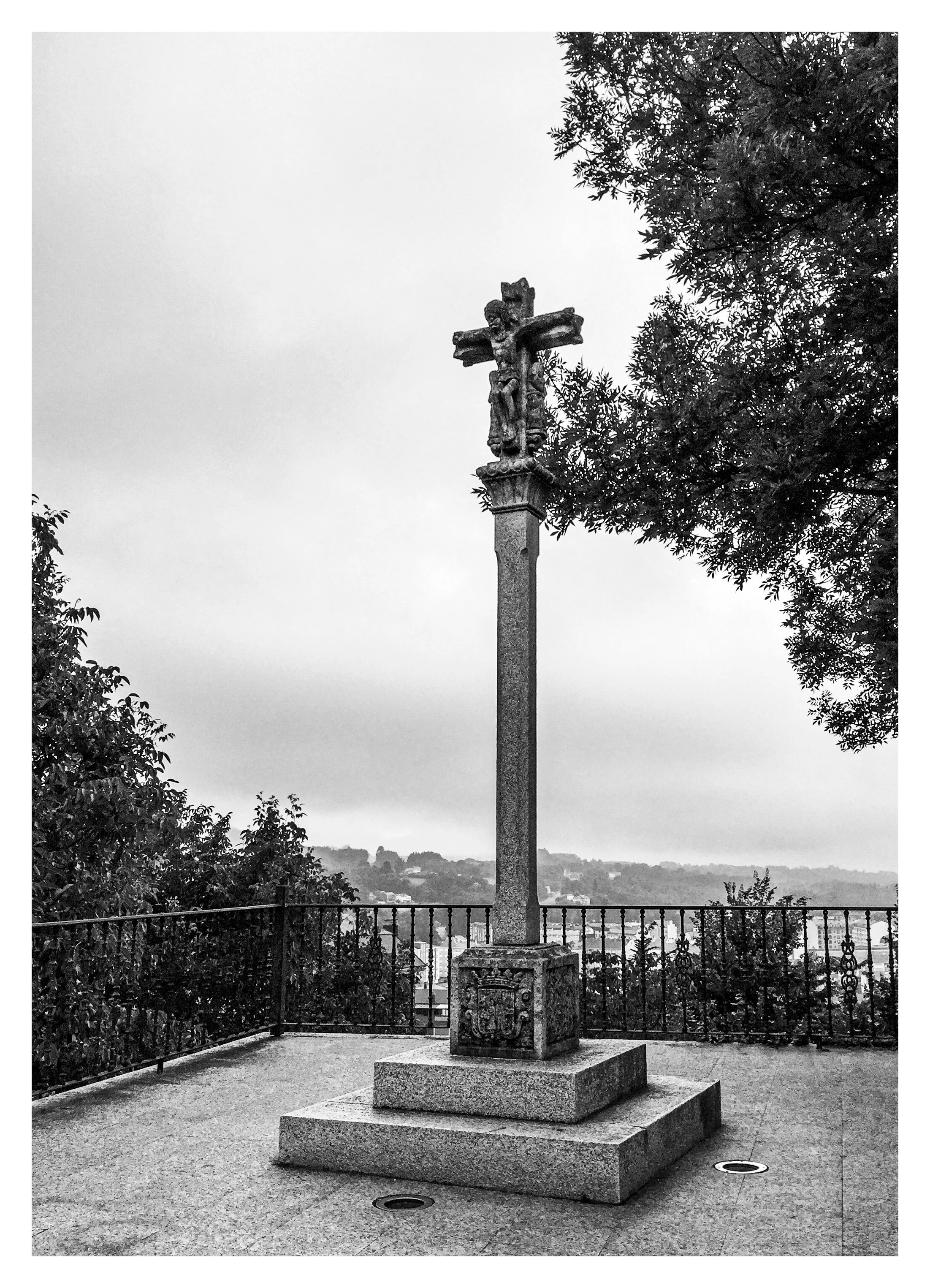 Black and white photograph of a stone Christian cross with a figure of Jesus Christ mounted on top, situated on a raised stone platform outdoors, with trees, a railing, and an overcast sky in the background.