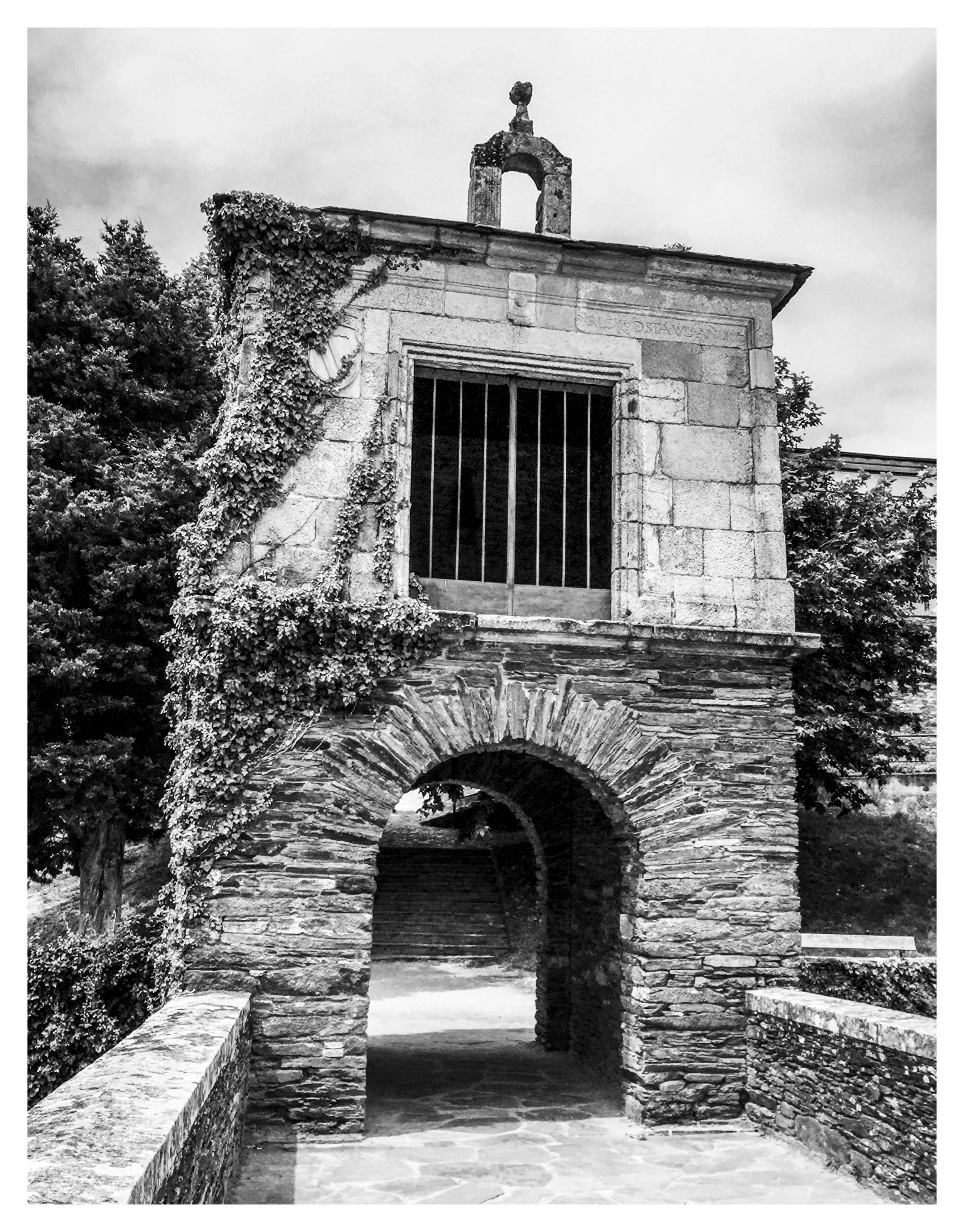Old stone gatehouse with an archway, barred window, ivy growing on the side, and a small bell tower on top, surrounded by trees.