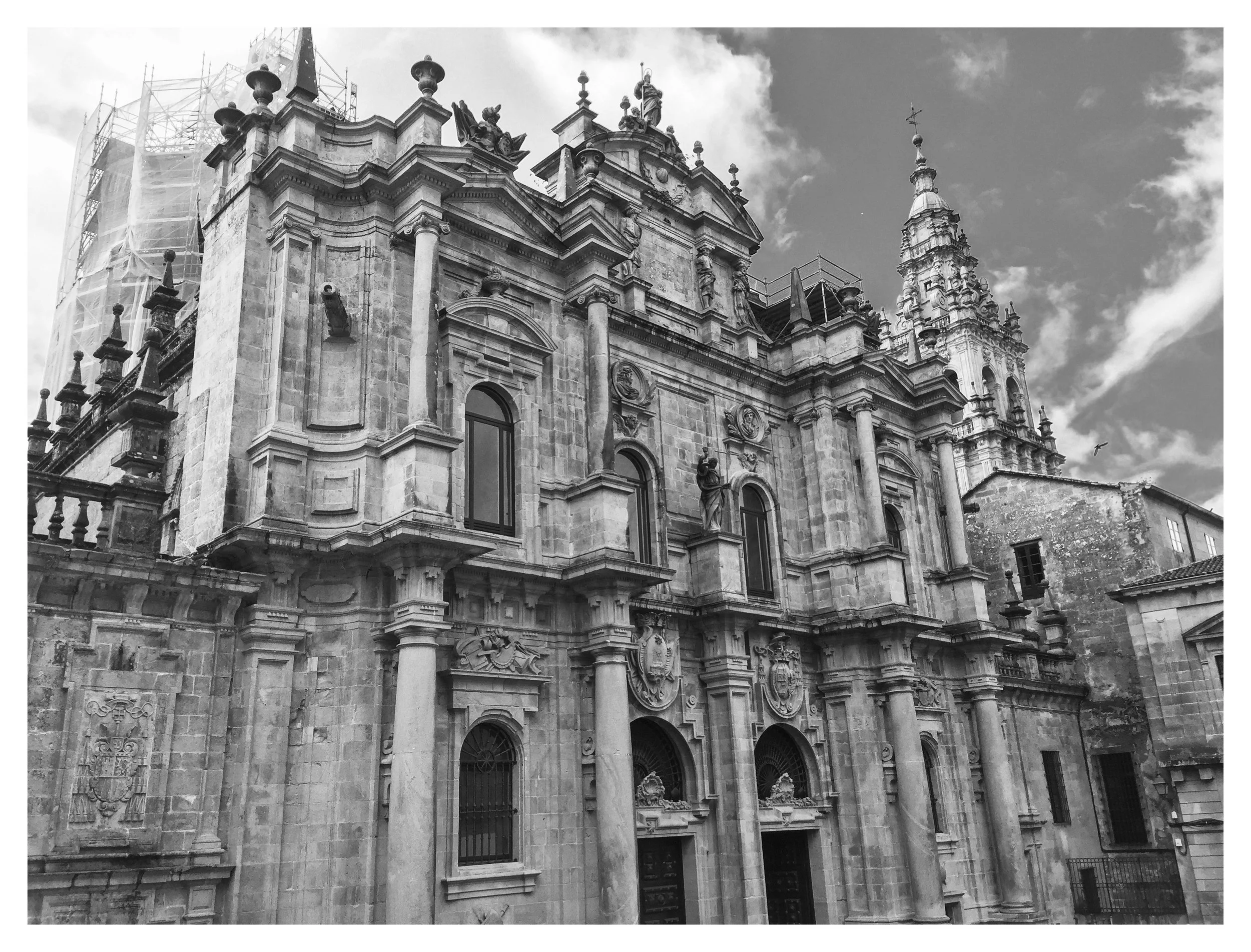 Black and white photo of a historic baroque-style church with ornate architectural details, statues, and decorative shields on its facade, with a cloudy sky in the background.