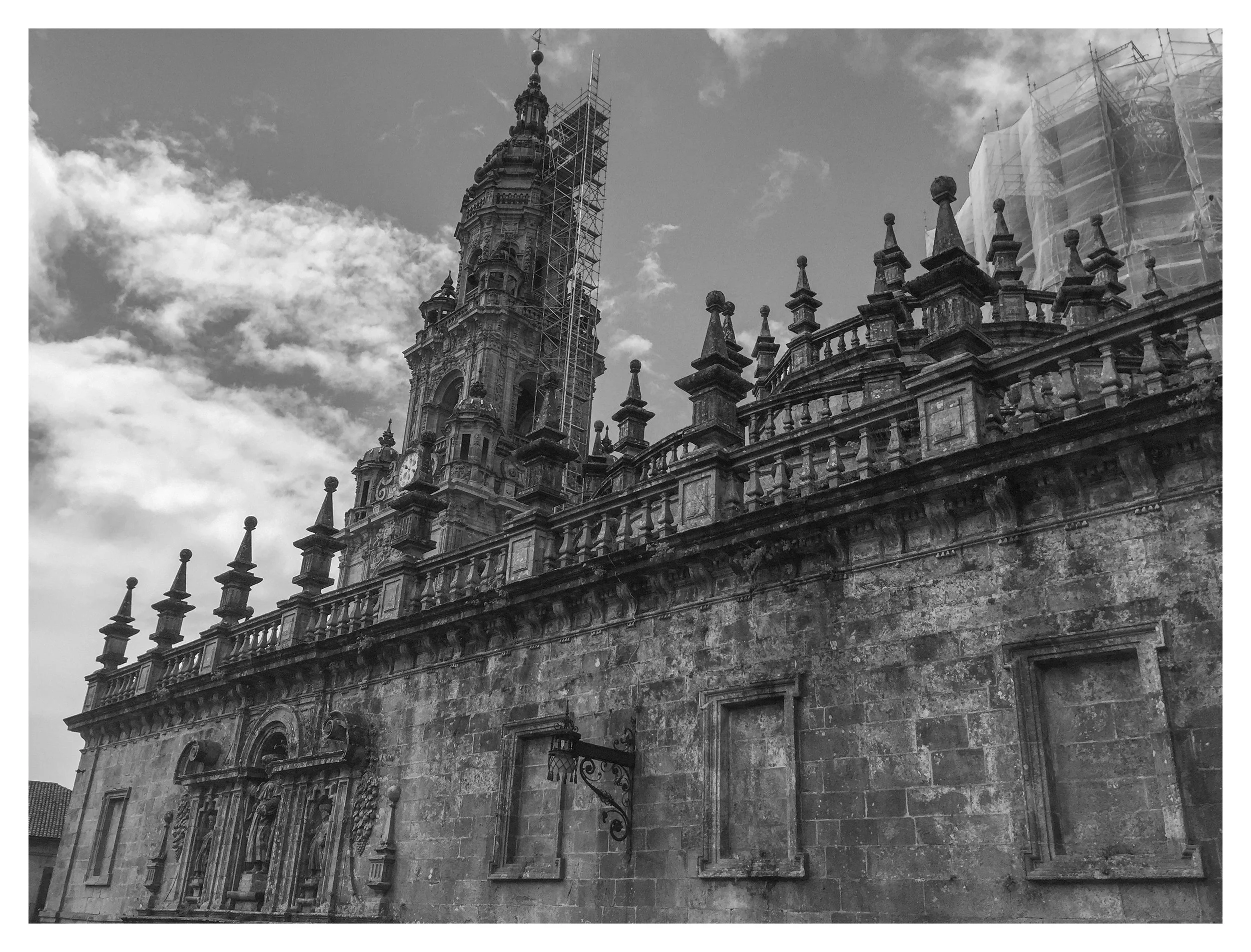 Black and white photo of an old historic building with ornate architectural details and a tall steeple, some scaffolding is visible on the steeple.