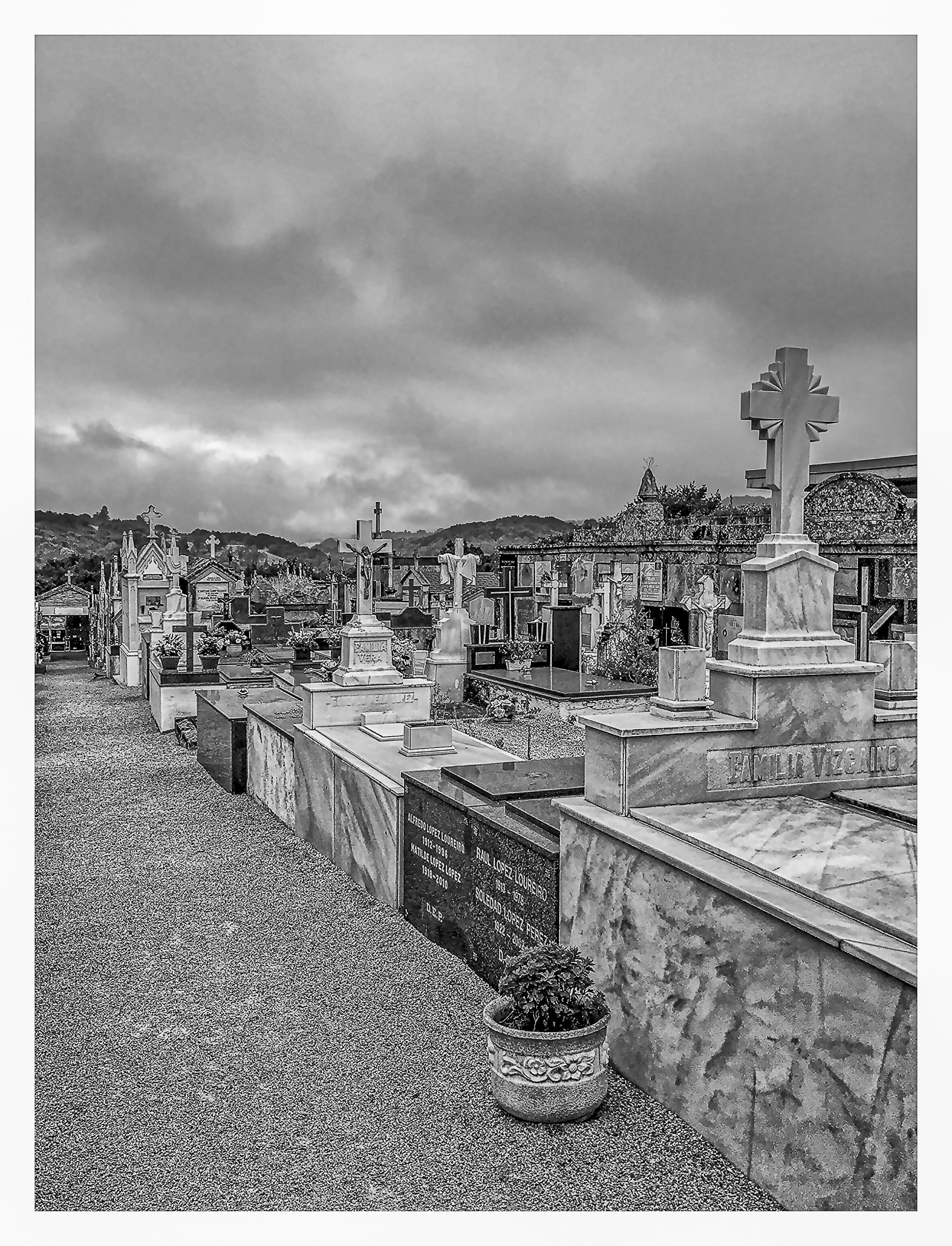 Black and white photo of a cemetery with numerous crosses and tombstones, some with flowers and plants, under a cloudy sky with hills in the background.