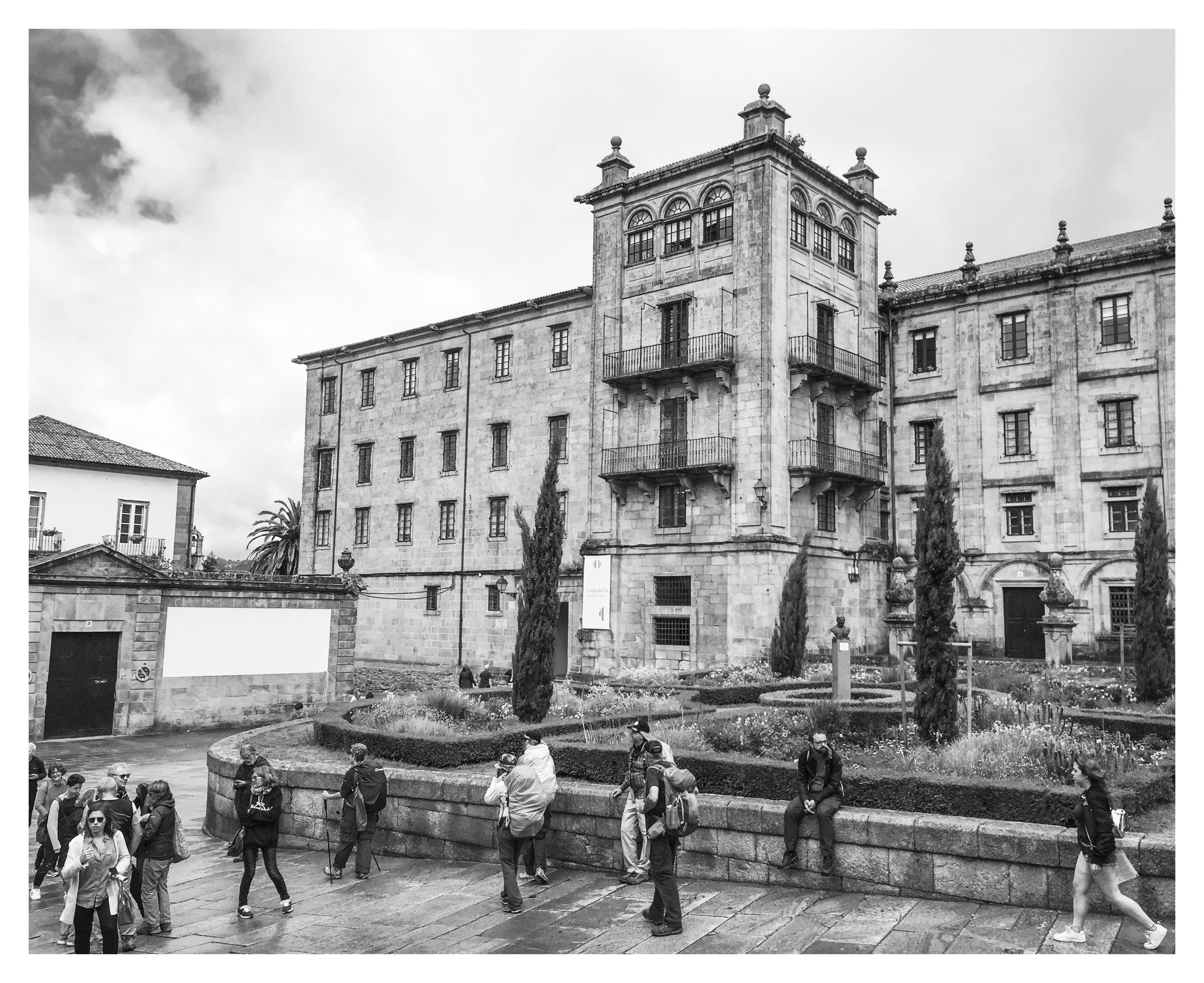 Historical stone building with multiple windows, balconies, and decorative columns, surrounded by a garden with tall cypress trees and people walking and sitting nearby.