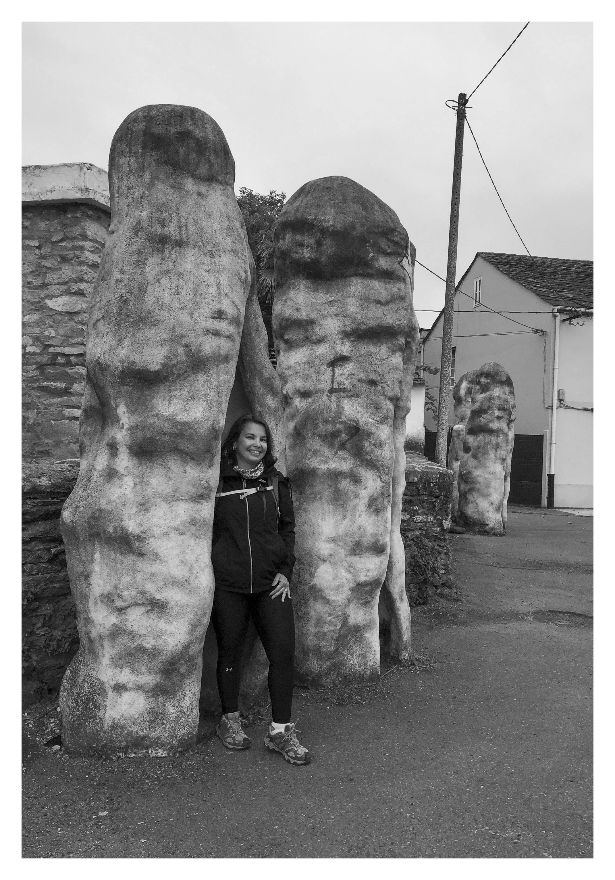 A woman standing smiling near a group of large, irregularly shaped sculptures made of stone or concrete, outdoors in a small town or village, in black and white.