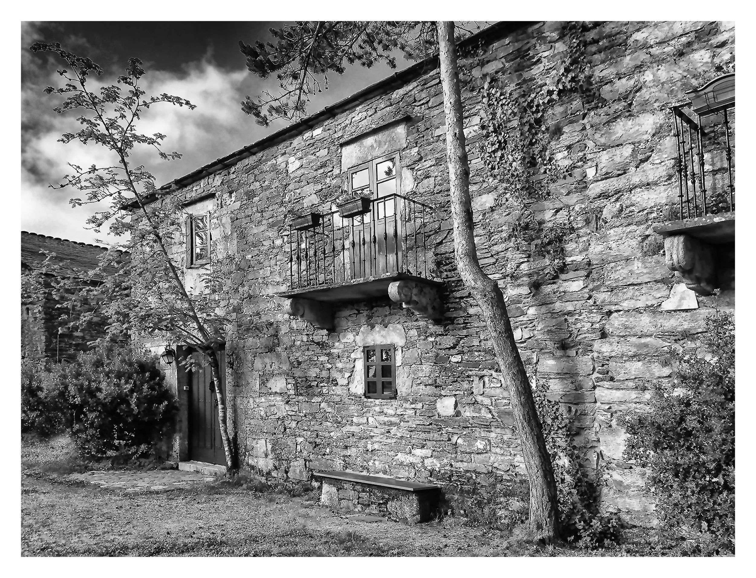 Black and white photo of a rustic stone building with small windows, a door, and small balconies with metal railings. There are trees and bushes around the building, and an overcast sky overhead.