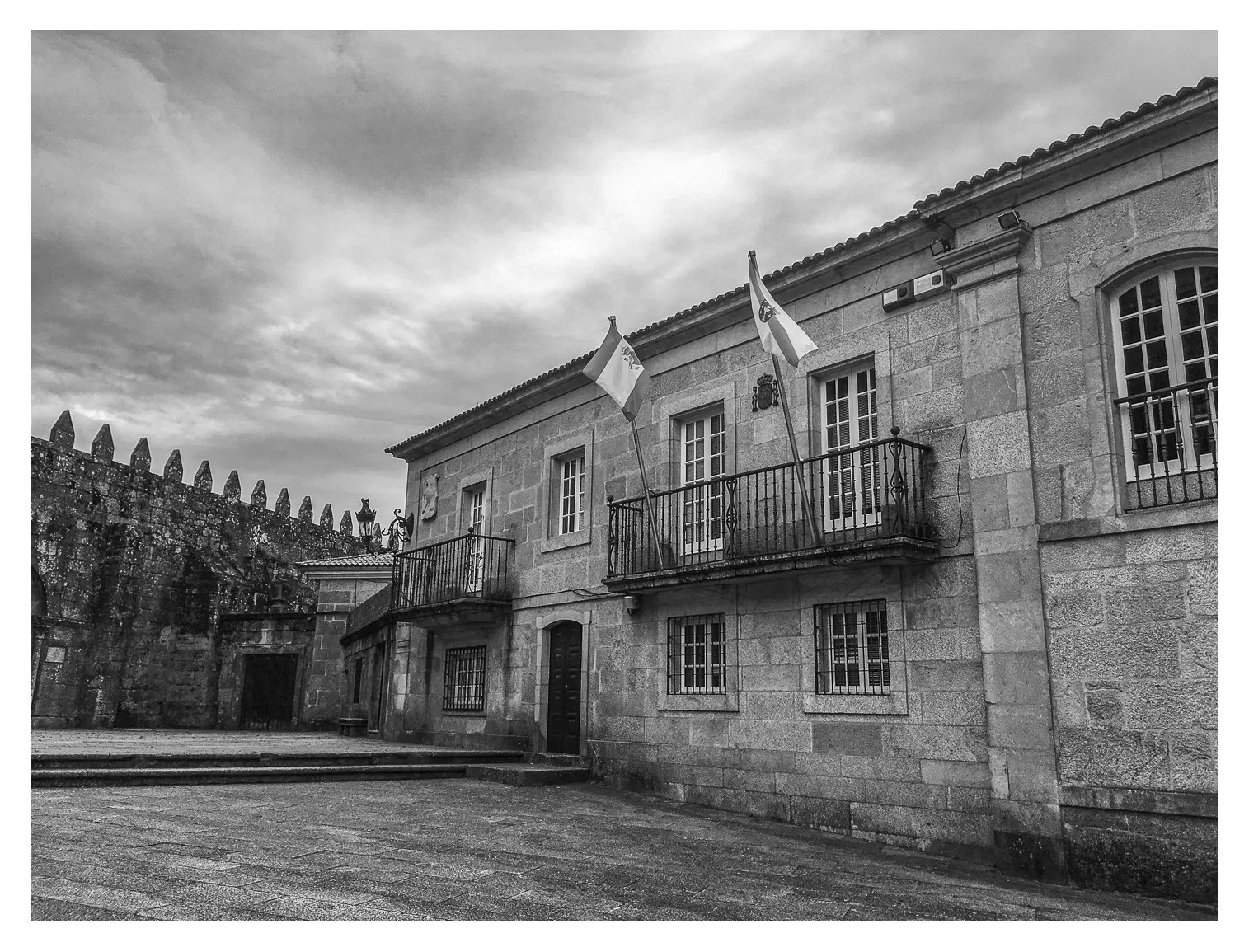 A stone building with a small balcony, two flags, and barred windows, situated near a stone wall with decorative battlements. The ground is wet, and the sky is overcast.