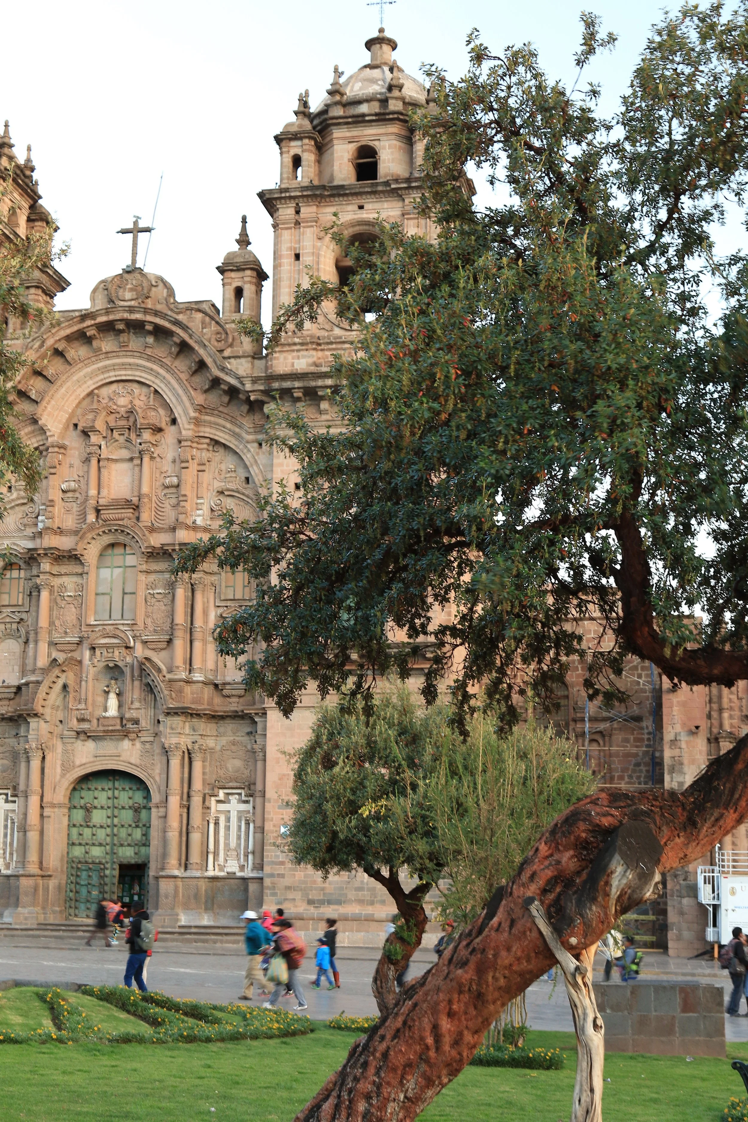 A historic stone church with intricate architectural details and a bell tower, partially obscured by large trees and greenery in a plaza with people walking by.