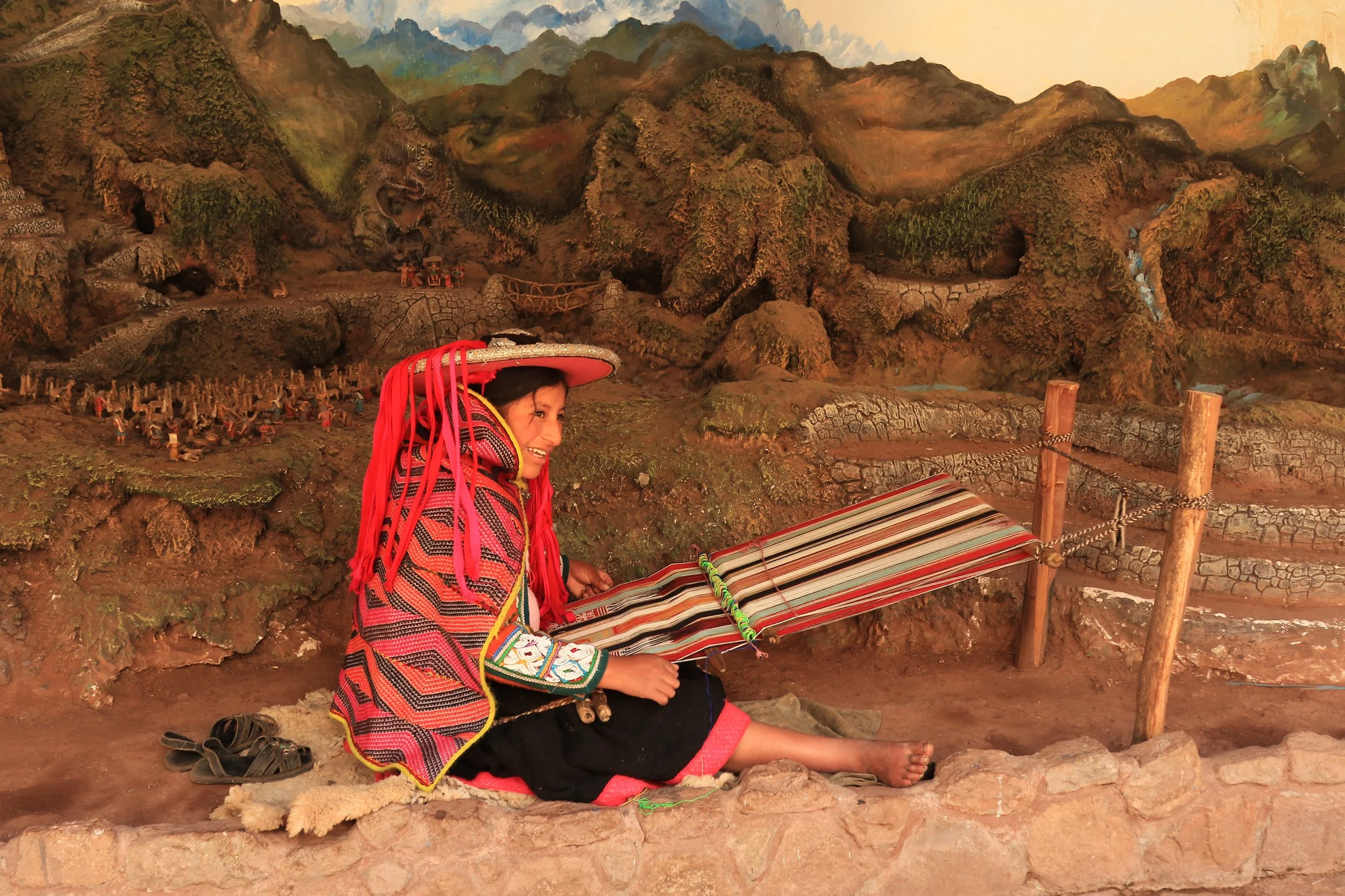 A woman from the indigenous Huichol community wearing traditional colorful clothing and a wide-brimmed hat, sitting on the ground while making a decorated yarn craft inside a cultural exhibit with a detailed mountain mural in the background.