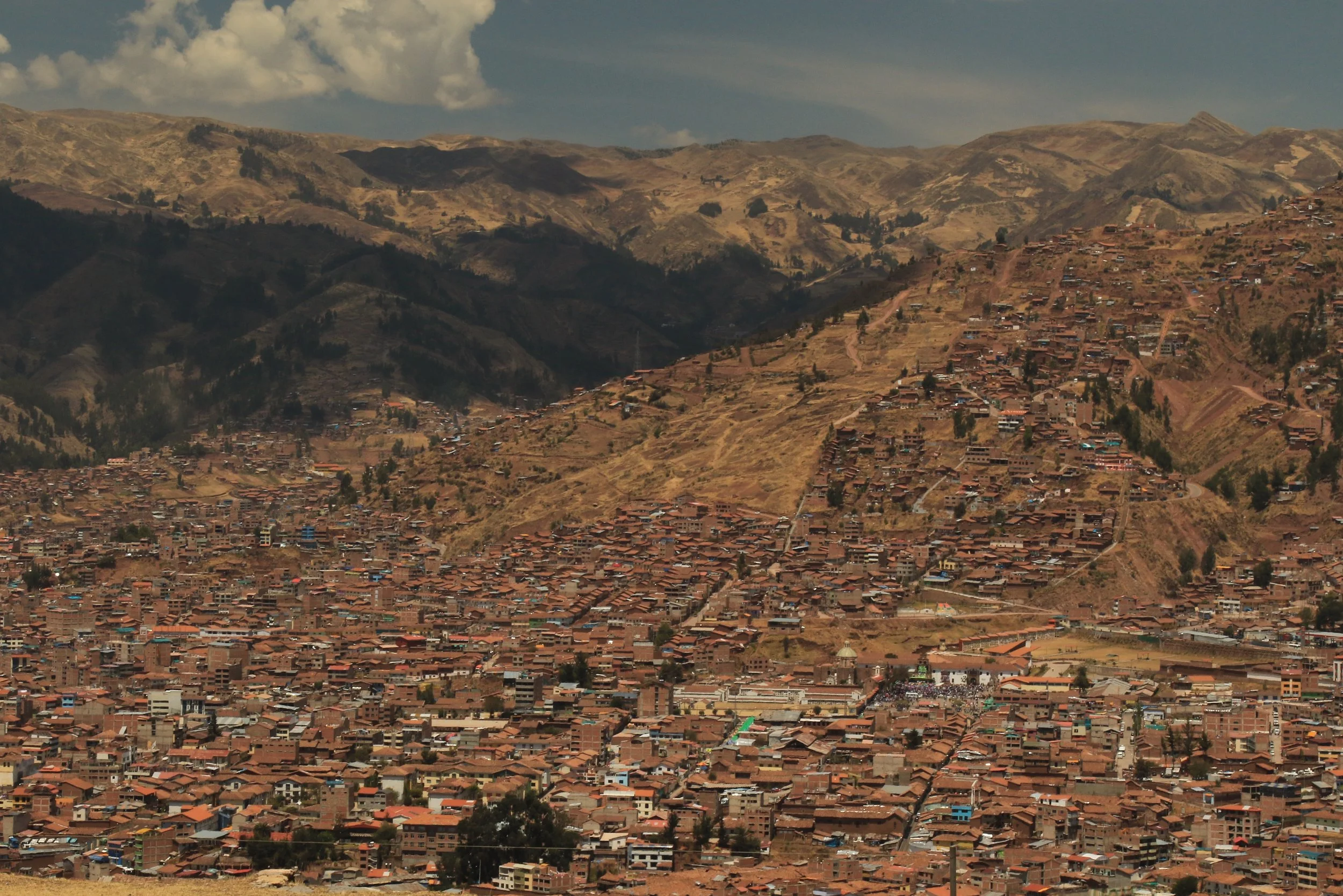 Panoramic view of a city built on hilly terrain with many brown rooftops, surrounded by mountainous landscape.