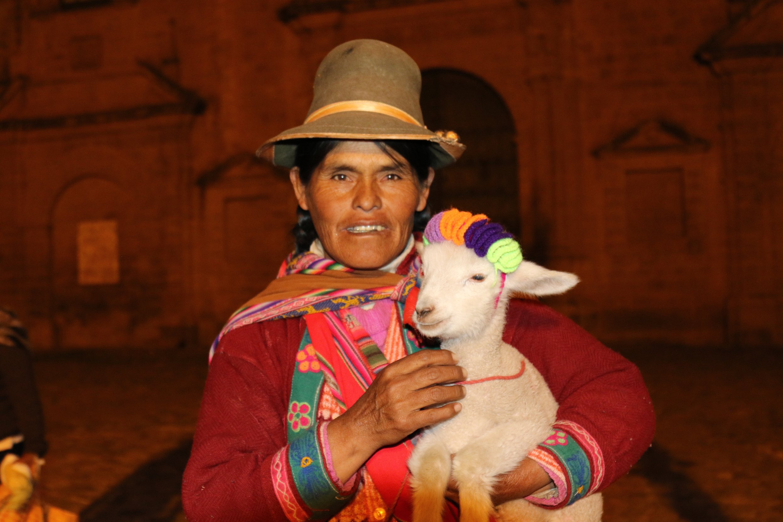 An indigenous woman in traditional colorful clothing holds a young white sheep with a rainbow-colored woolly headband. The woman is wearing a wide-brimmed hat and a red sweater with detailed embroidery, standing in front of an ancient brick building 